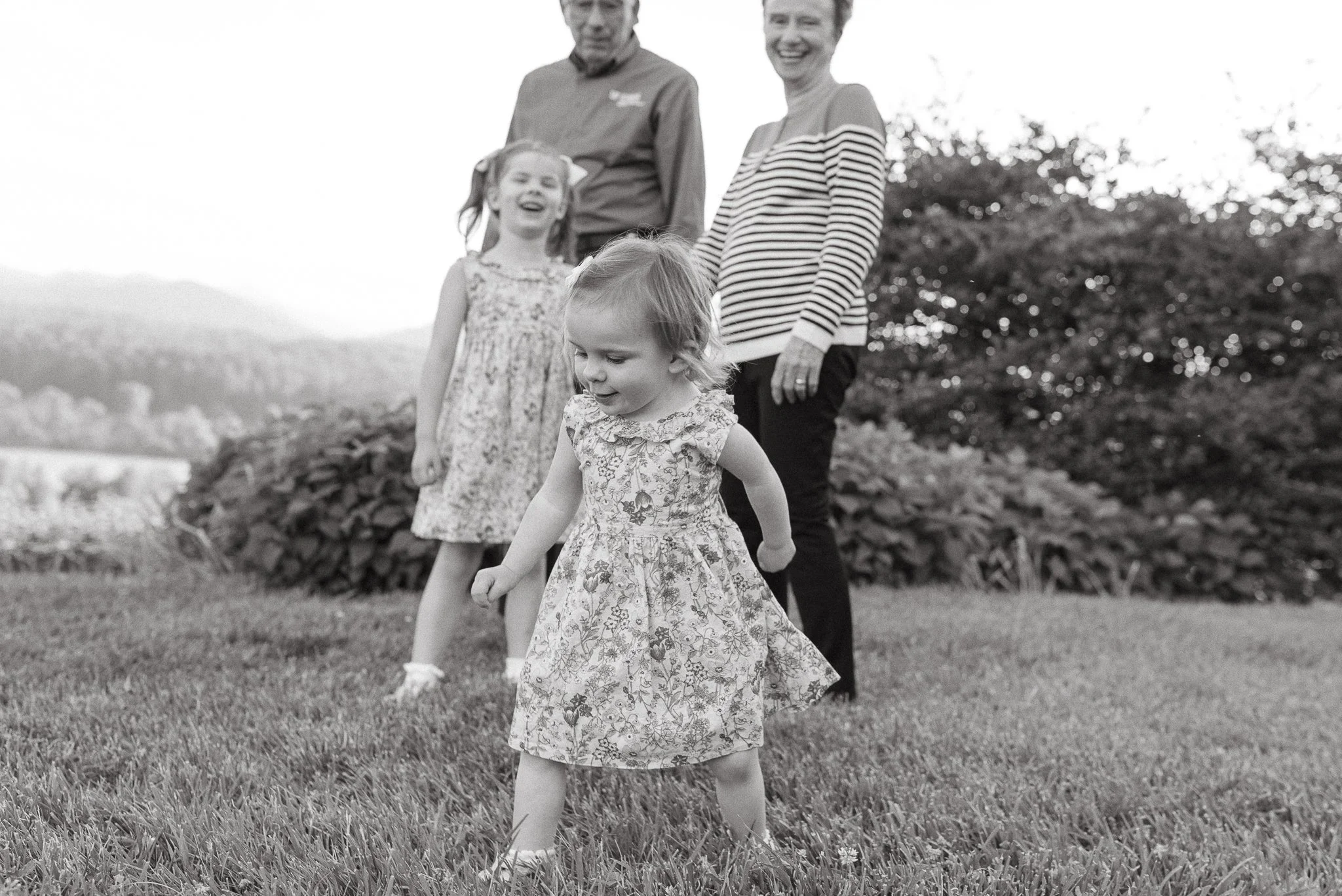 A group of four people, including three women and one girl, standing outdoors on grass with a blurred natural background. The girl in the foreground is looking down, while the others behind her are smiling and looking at the camera.