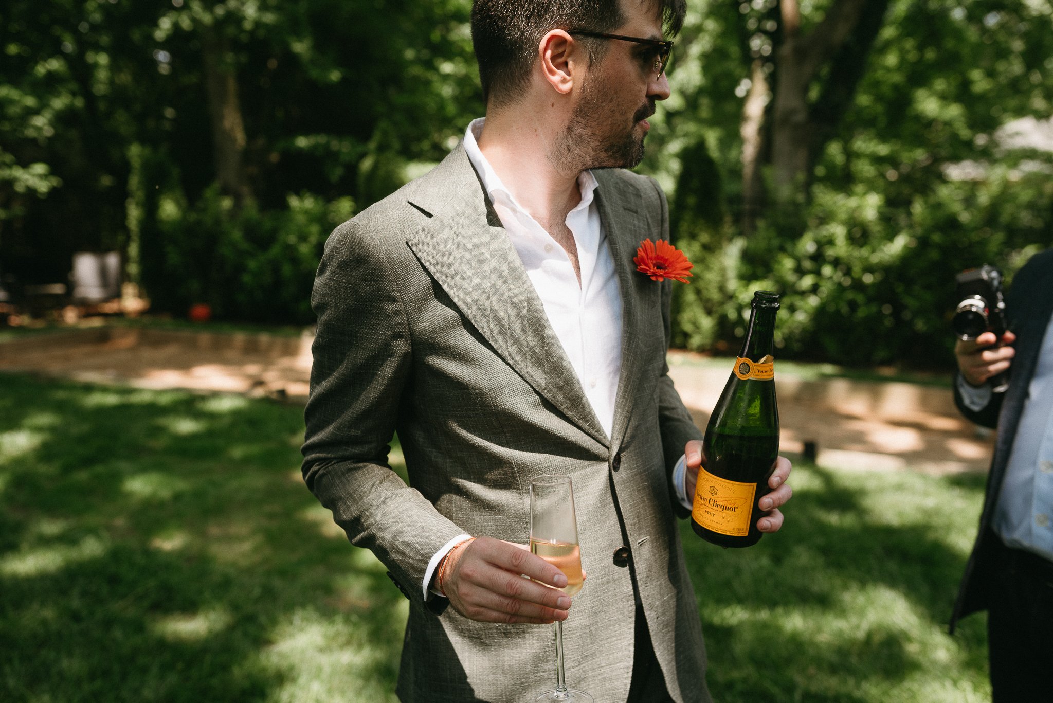 A man in a gray suit with a flower in the lapel holds a champagne bottle and glass at an outdoor event, with greenery in the background.