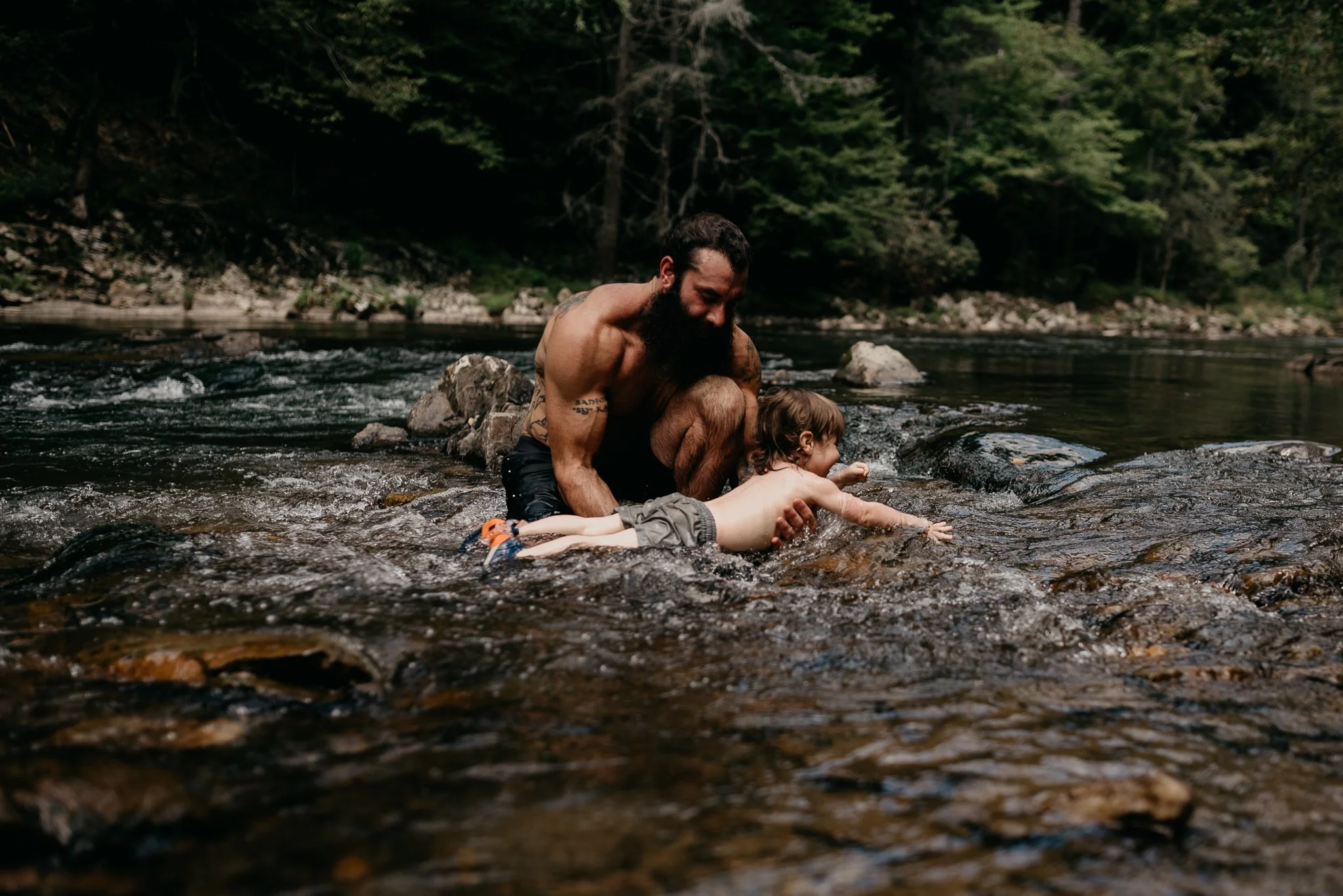 A shirtless man with a beard helping a young child swim in a river surrounded by trees and rocks.