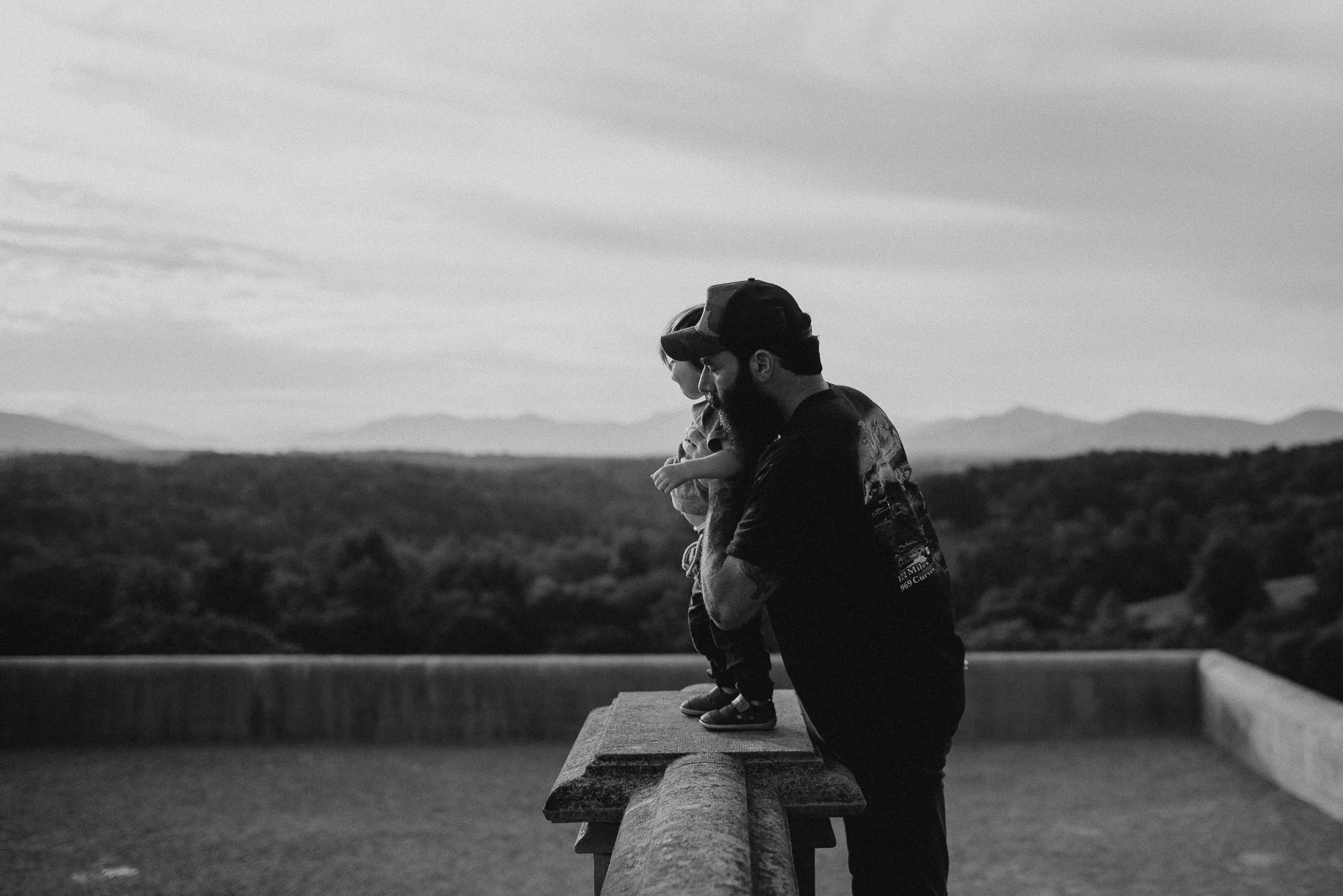 A man and a young girl are kneeling together on a low wall outdoors, with a scenic landscape of hills or mountains in the background. They are looking downward, possibly at something on the ground or in the water, and seem to be sharing a quiet momen