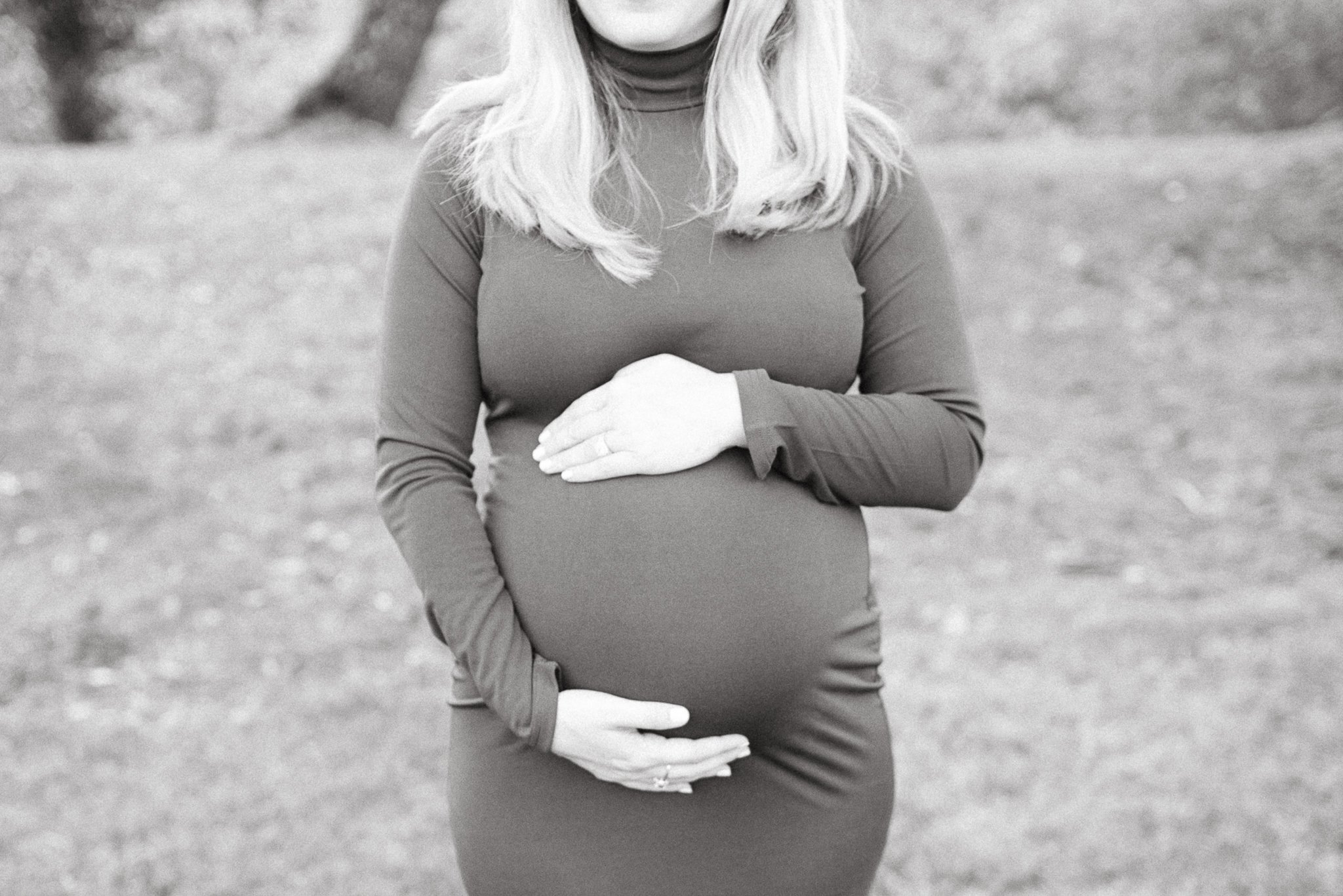 A woman in a long sleeve dress with blonde hair, holding her pregnant belly with both hands, outdoors on a gravel or sand surface.
