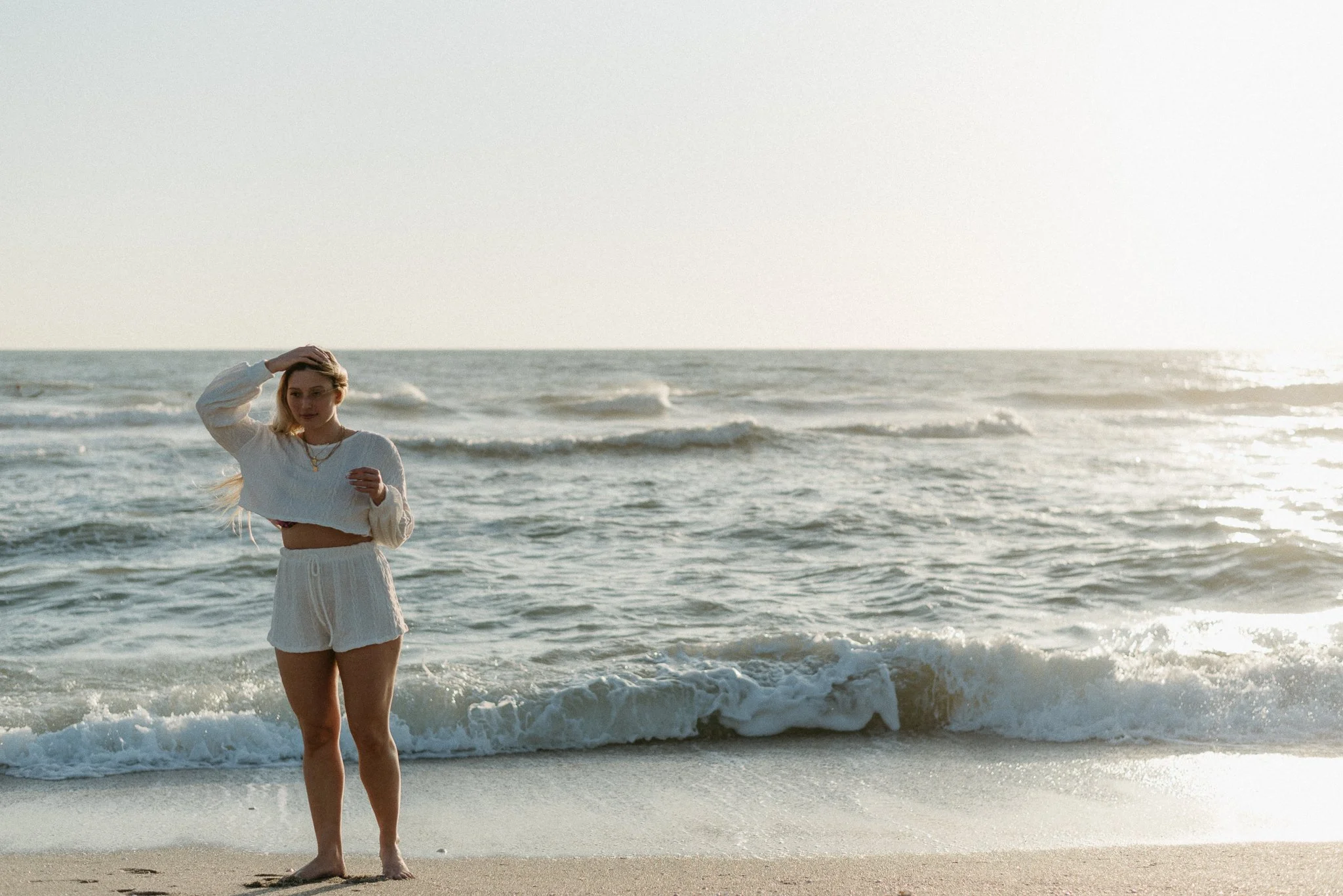 A woman standing barefoot on a sandy beach with ocean waves behind her, wearing a white sweater and matching shorts, with one hand on her head and the other near her chest, during sunset.