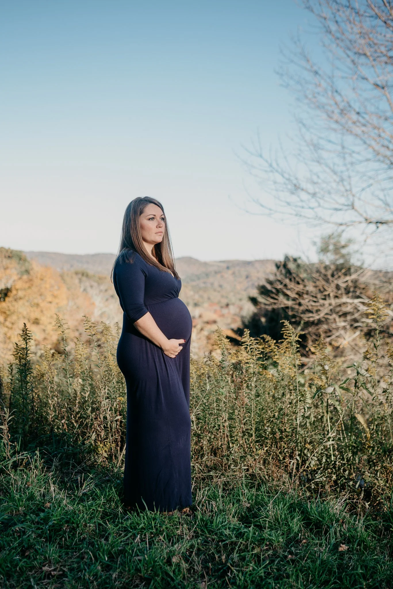 A pregnant woman in a navy blue dress stands outdoors in a field with tall grass and plants, with hills and a clear blue sky in the background.