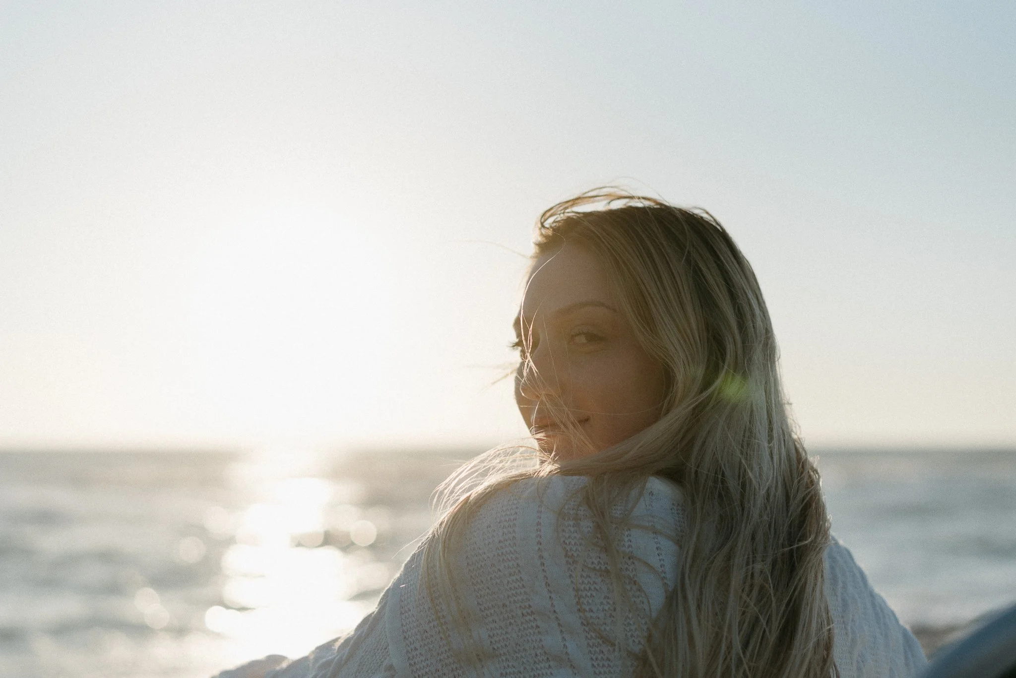 A woman with long blonde hair with waves, smiling gently at the camera, is sitting outdoors near a body of water during sunset or sunrise, with sunlight reflecting off the water and a clear sky.
