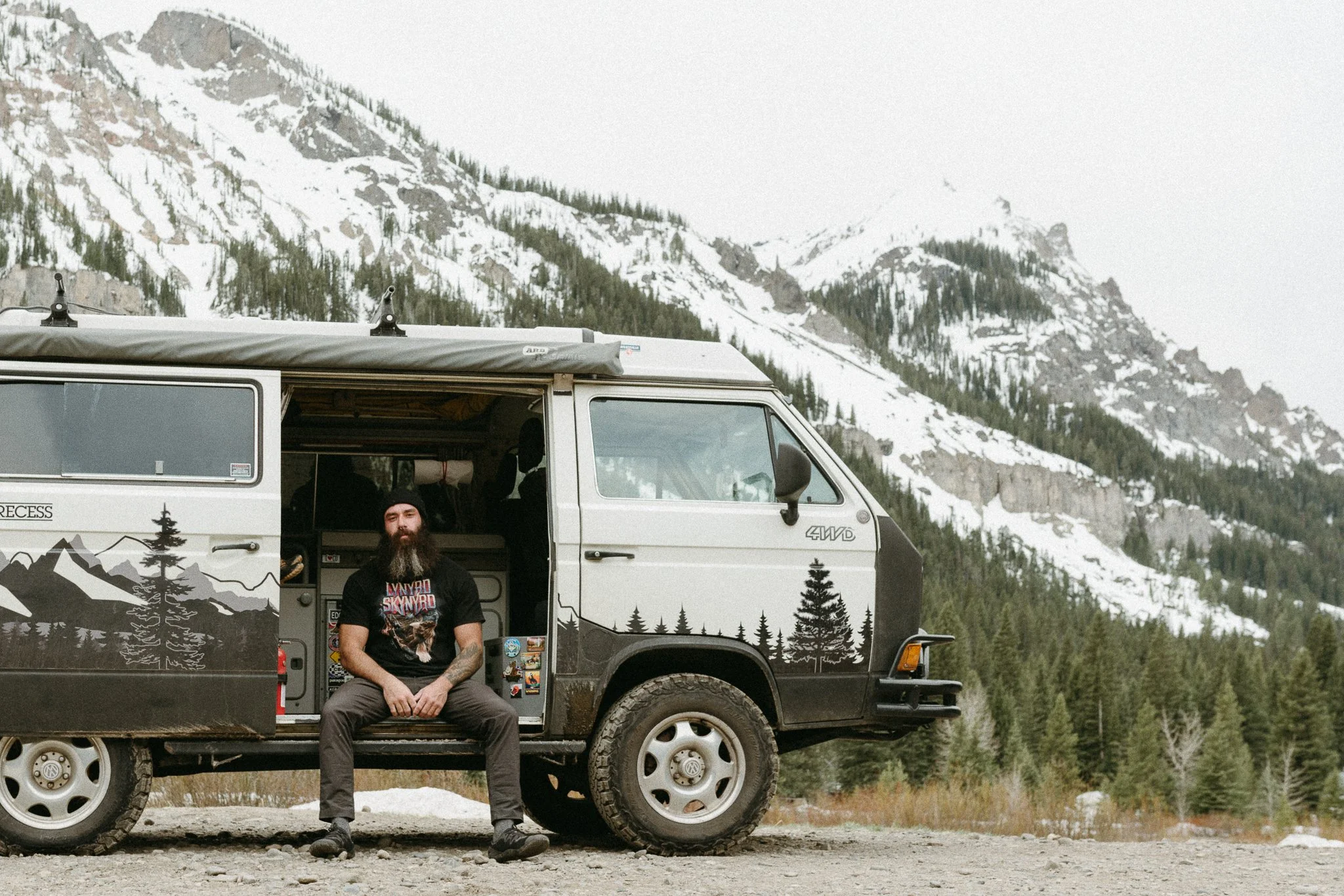 A man with a beard and tattooed arms sitting on the running board of a black off-road vehicle with mountain scenery and snow in the background.