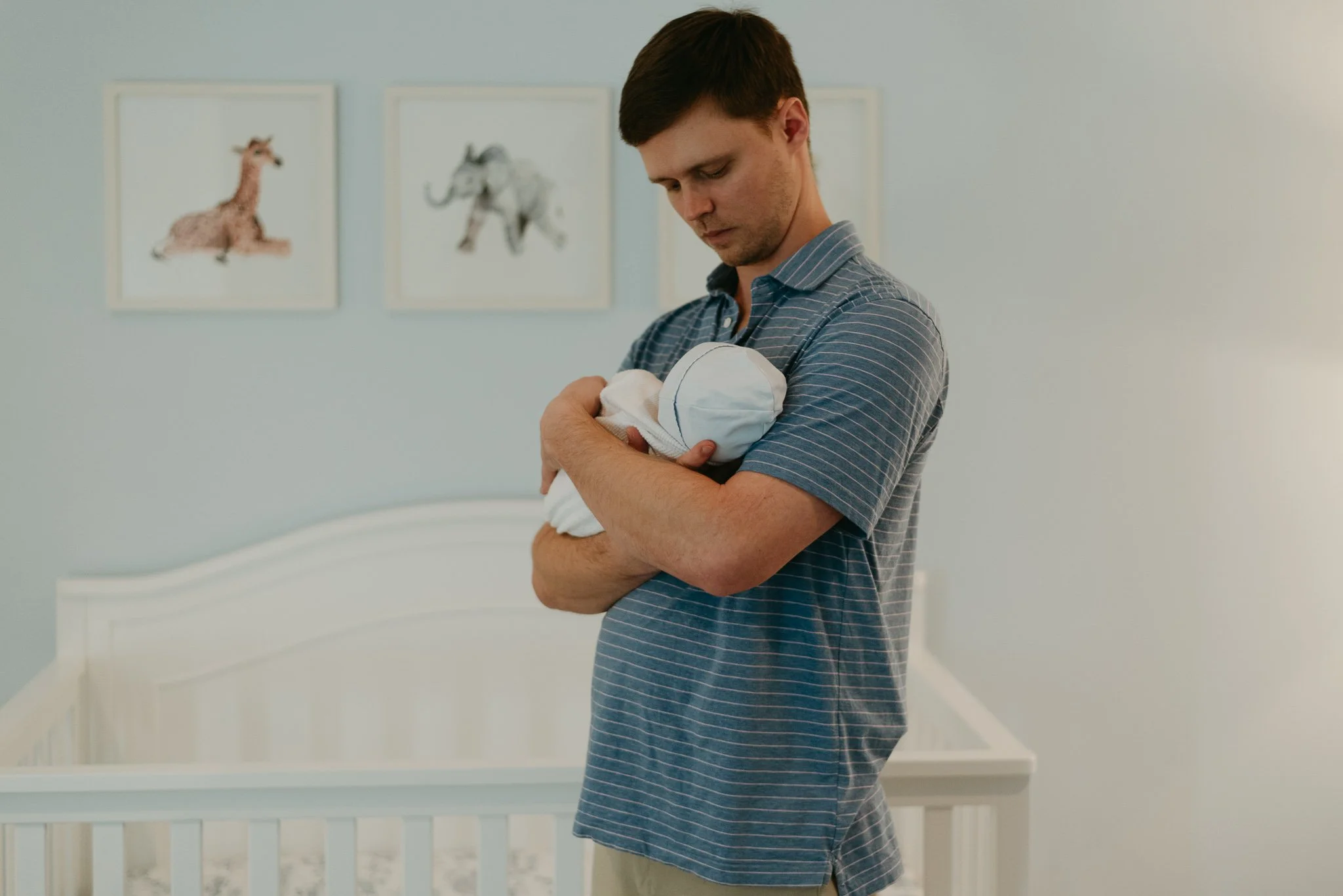 A young man holding a baby wrapped in a blanket, standing in a white nursery with picture frames of animals on the wall.
