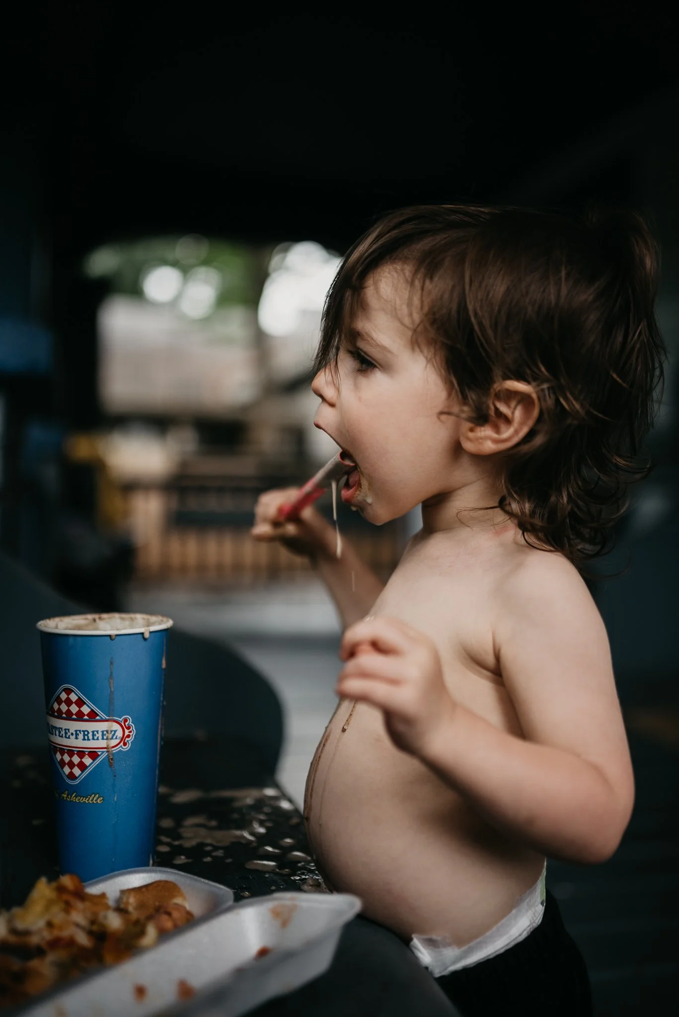A young shirtless boy with brown hair is eating food using a fork at a table with a blue icee cup and food in takeout containers.