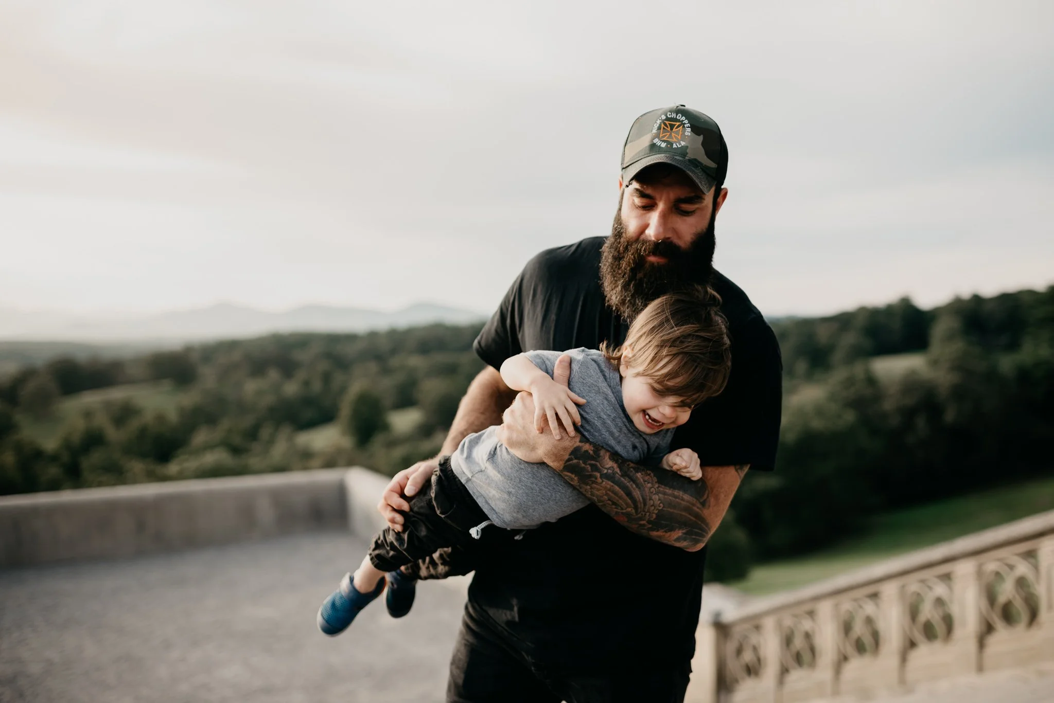 A man with a beard wearing a cap and black t-shirt is holding and lifting a young boy with blonde hair in a gray shirt and blue shoes in an outdoor setting with a cloudy sky and distant green hills.
