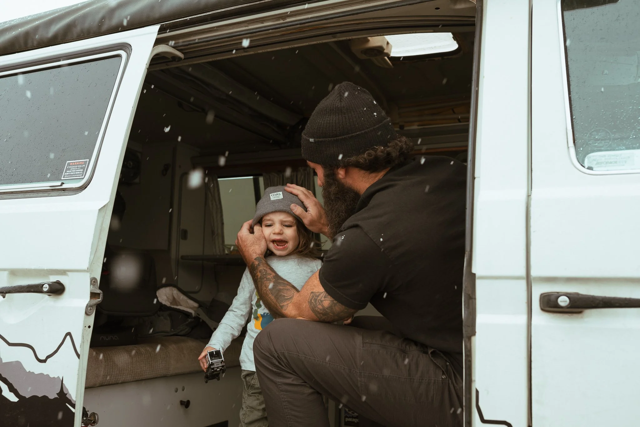 A man with tattoos holding a young girl with a toy truck inside a white camper van in snowy weather.