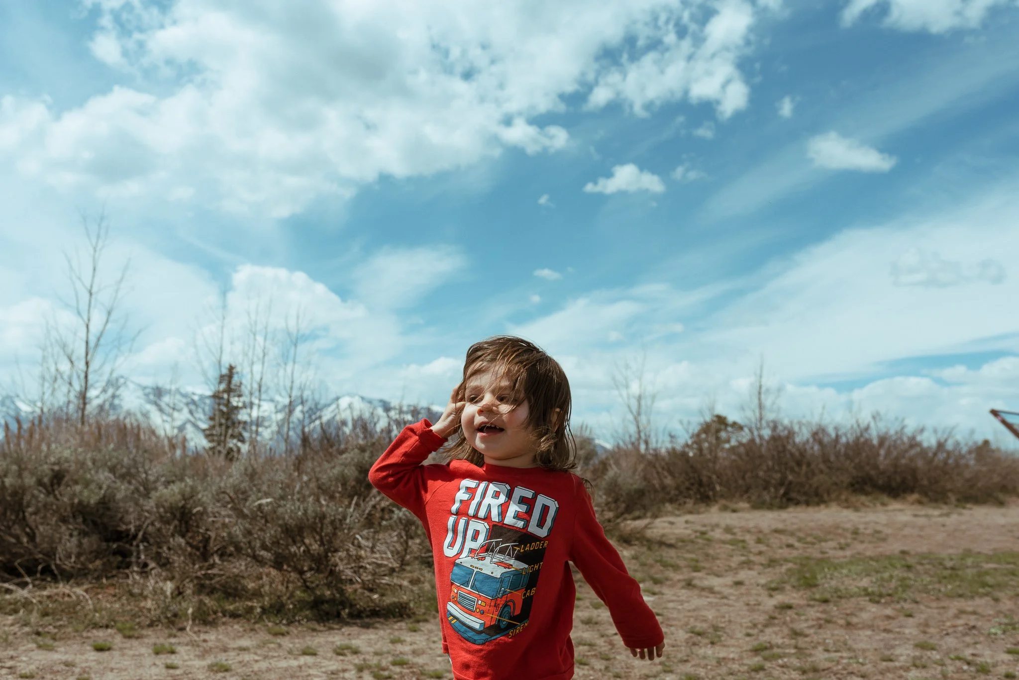 A young child with brown hair wearing a red fire truck shirt, smiling and touching their ear while standing outdoors under a partly cloudy sky, with leafless trees and mountain peaks in the background.