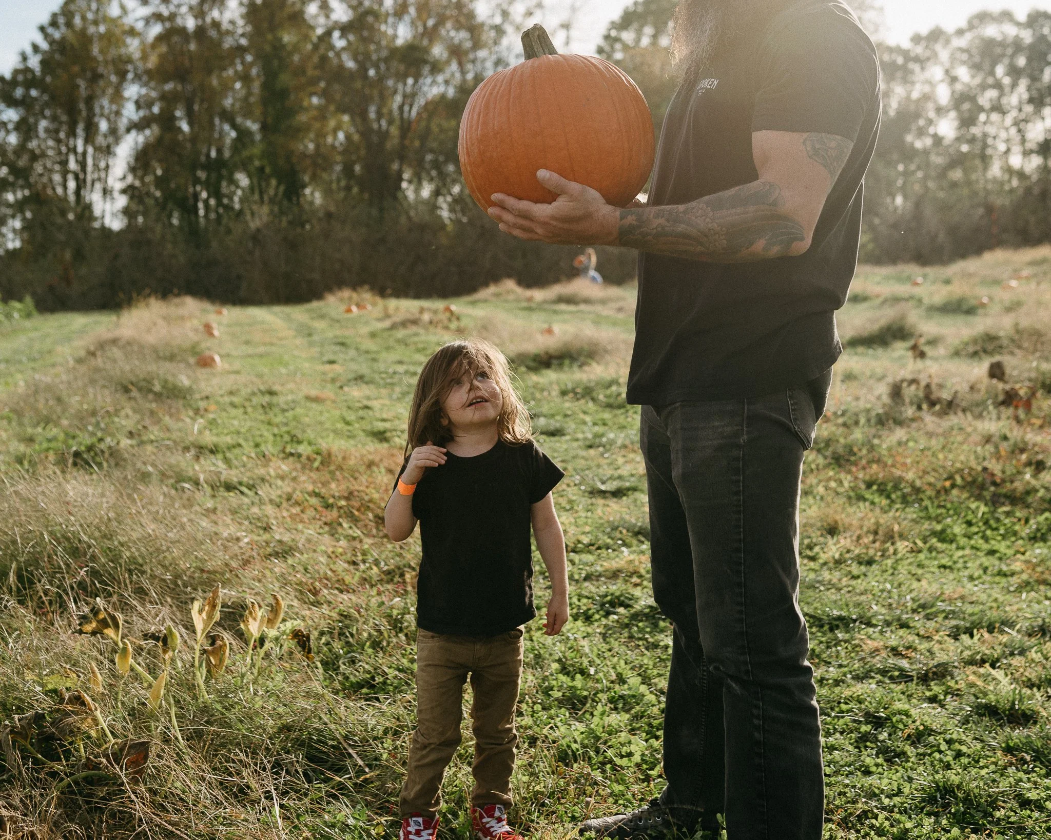 A man holding a pumpkin while a child looks up at him in a grassy field during daytime.