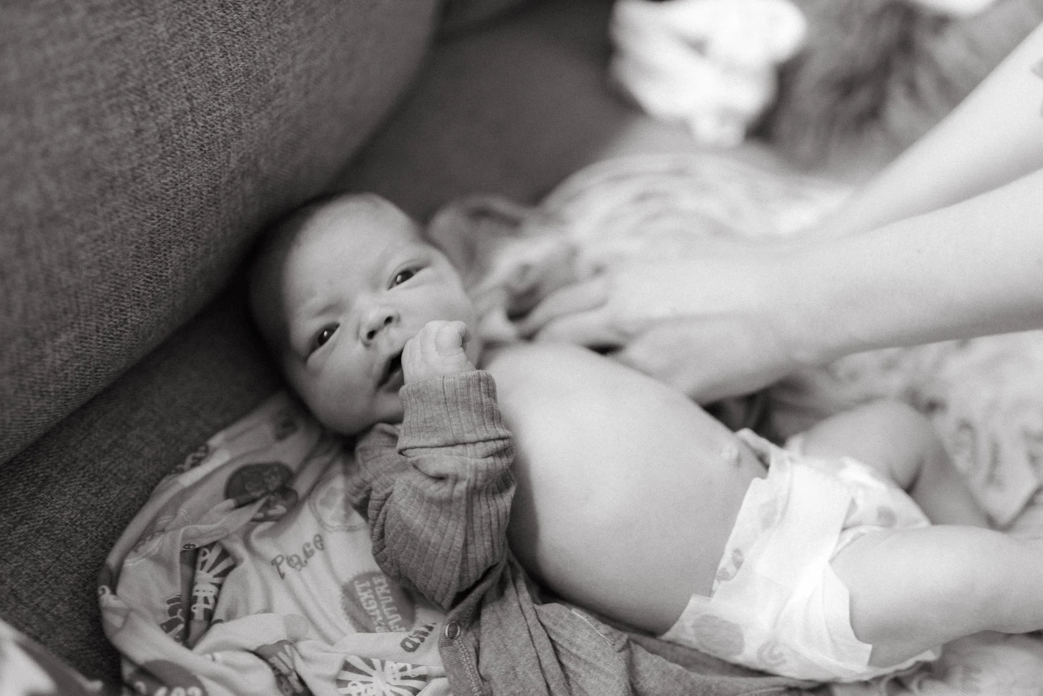 A baby lying on a blanket in a crib, looking up with one hand near their face, in a grayscale photograph.