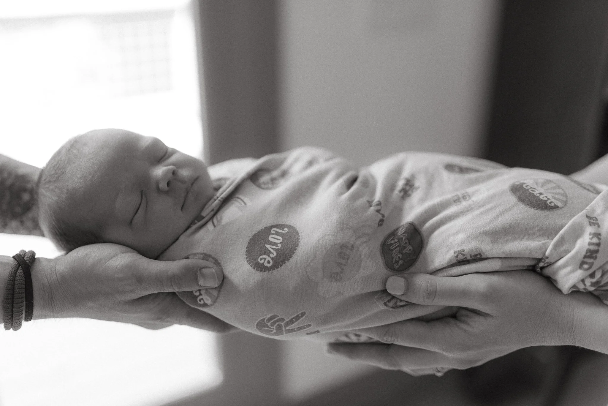 A newborn baby sleeping peacefully, being gently held by an adult's hand, with the focus on the baby's face and the adult's hand supporting the baby, in a monochrome photo.