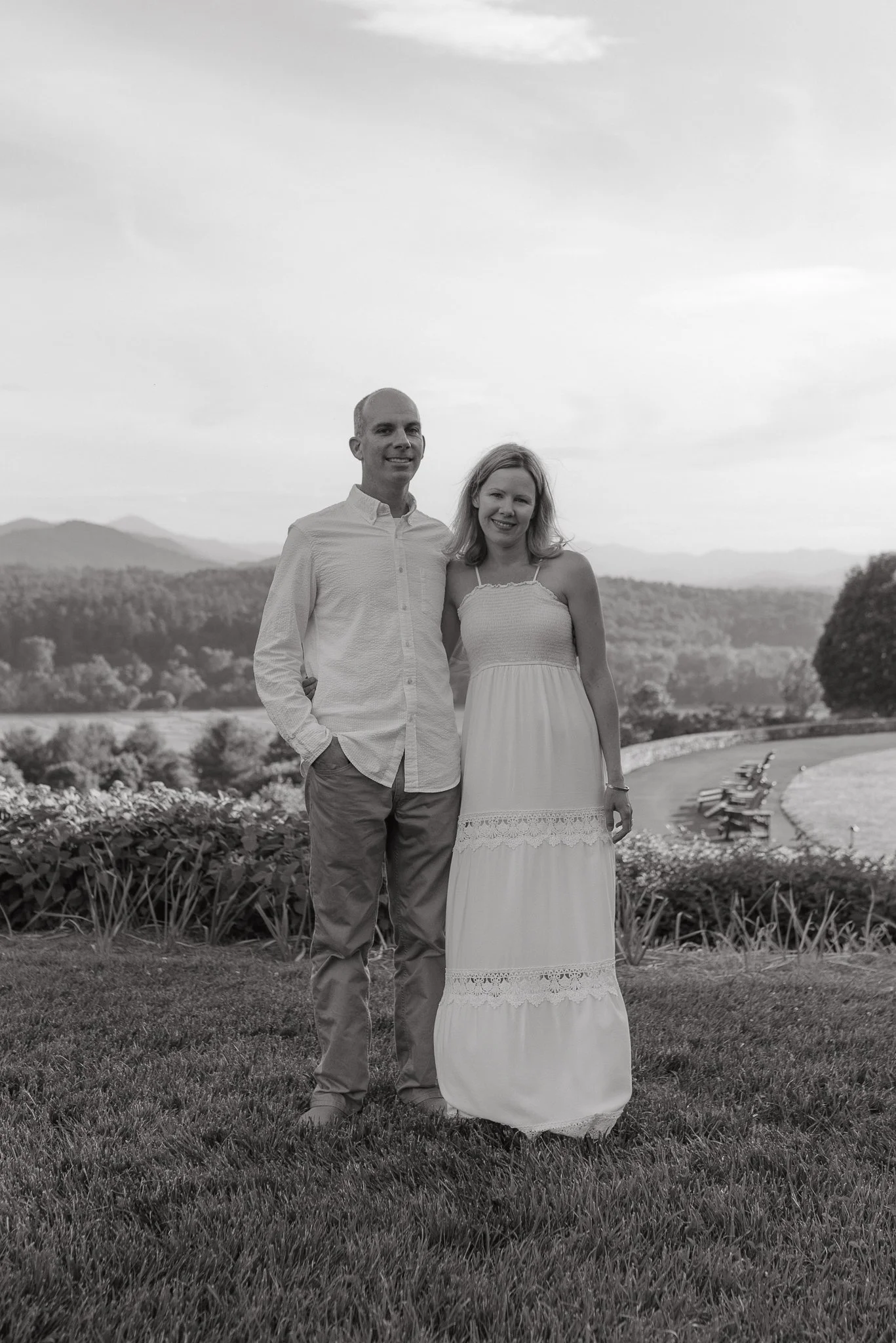 A black and white photo of a smiling couple standing outdoors, with mountains and trees in the background. The man is wearing a light-colored button-up shirt and khakis, and the woman is in a long, strapless dress.