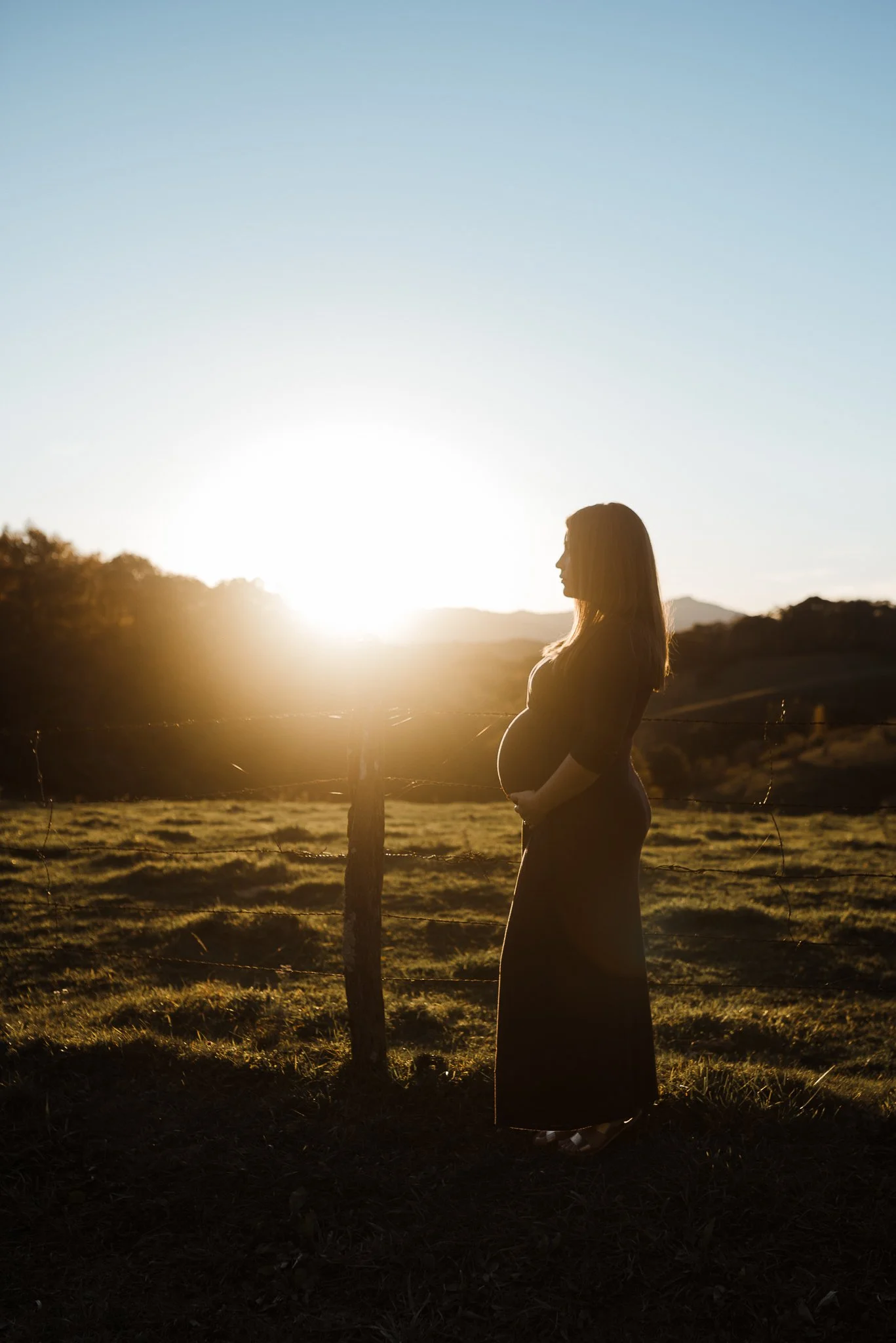 A pregnant woman standing outdoors near a wooden fence at sunset or sunrise.