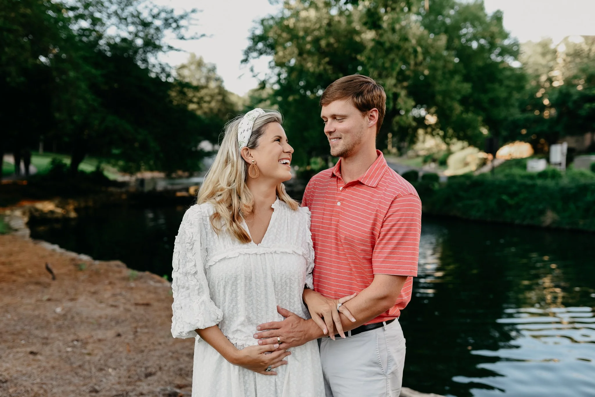 A couple stands close together in a park near a river, smiling and looking into each other's eyes. The woman has long wavy hair, is wearing a white dress, and a headband, while the man has short brown hair, is wearing a red striped shirt, and white pants. They are surrounded by green trees and natural scenery.