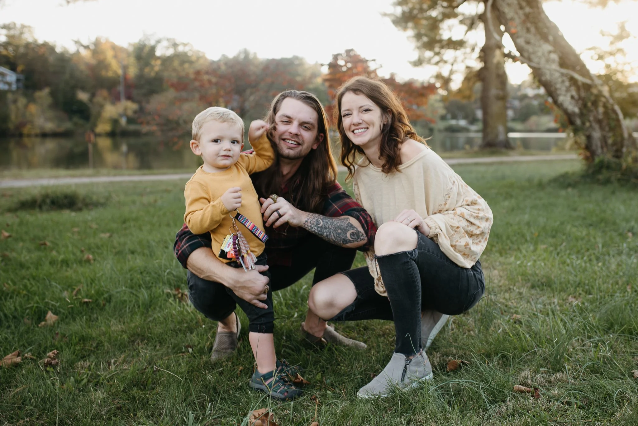 A family of three, a man, woman, and young boy, outdoors in a park with green grass, trees with fall foliage, and a lake in the background. The man has long hair and tattoos, the woman has curly hair, and the boy has blonde hair and is wearing a yell