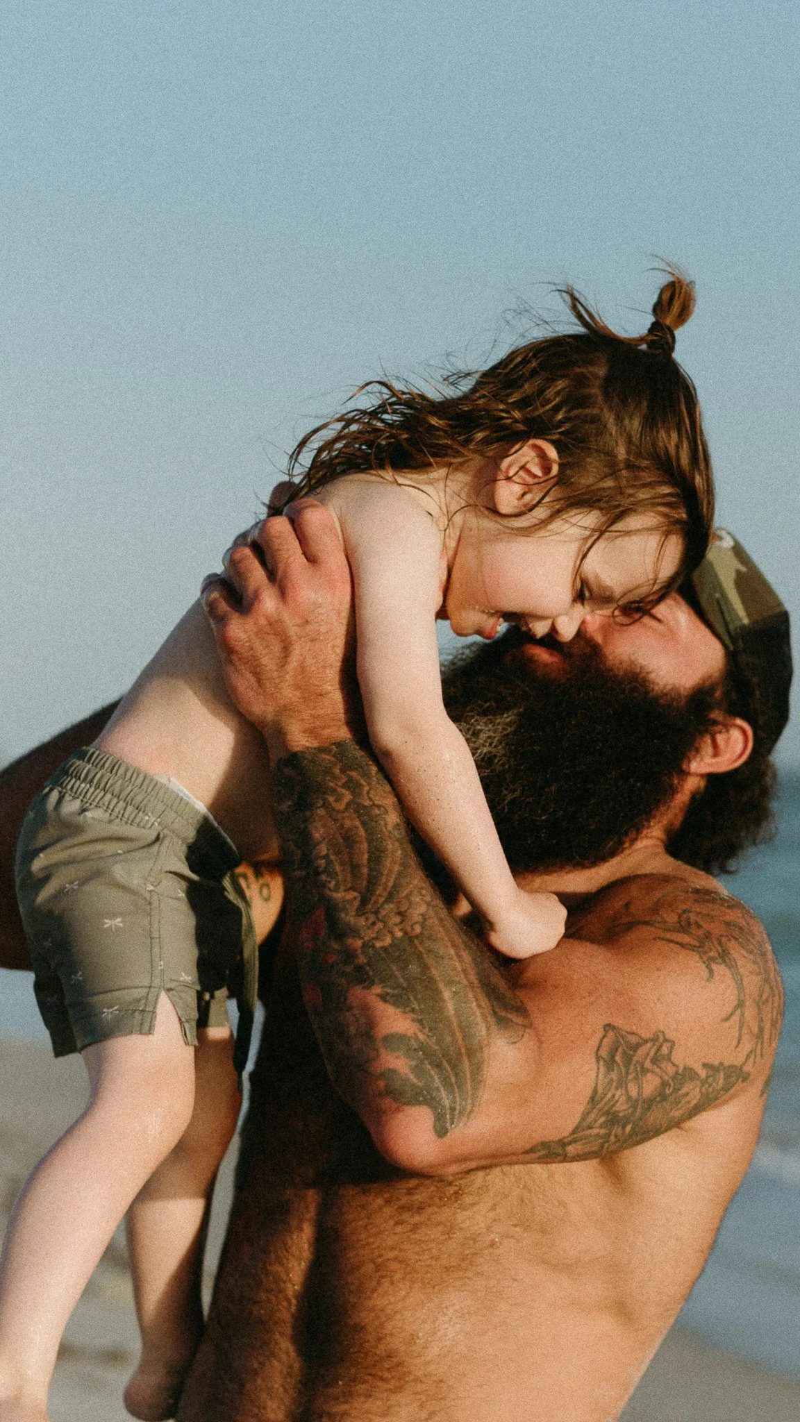 A shirtless man with tattoos holding a young girl on his shoulders, both smiling and enjoying time outdoors during the day.
