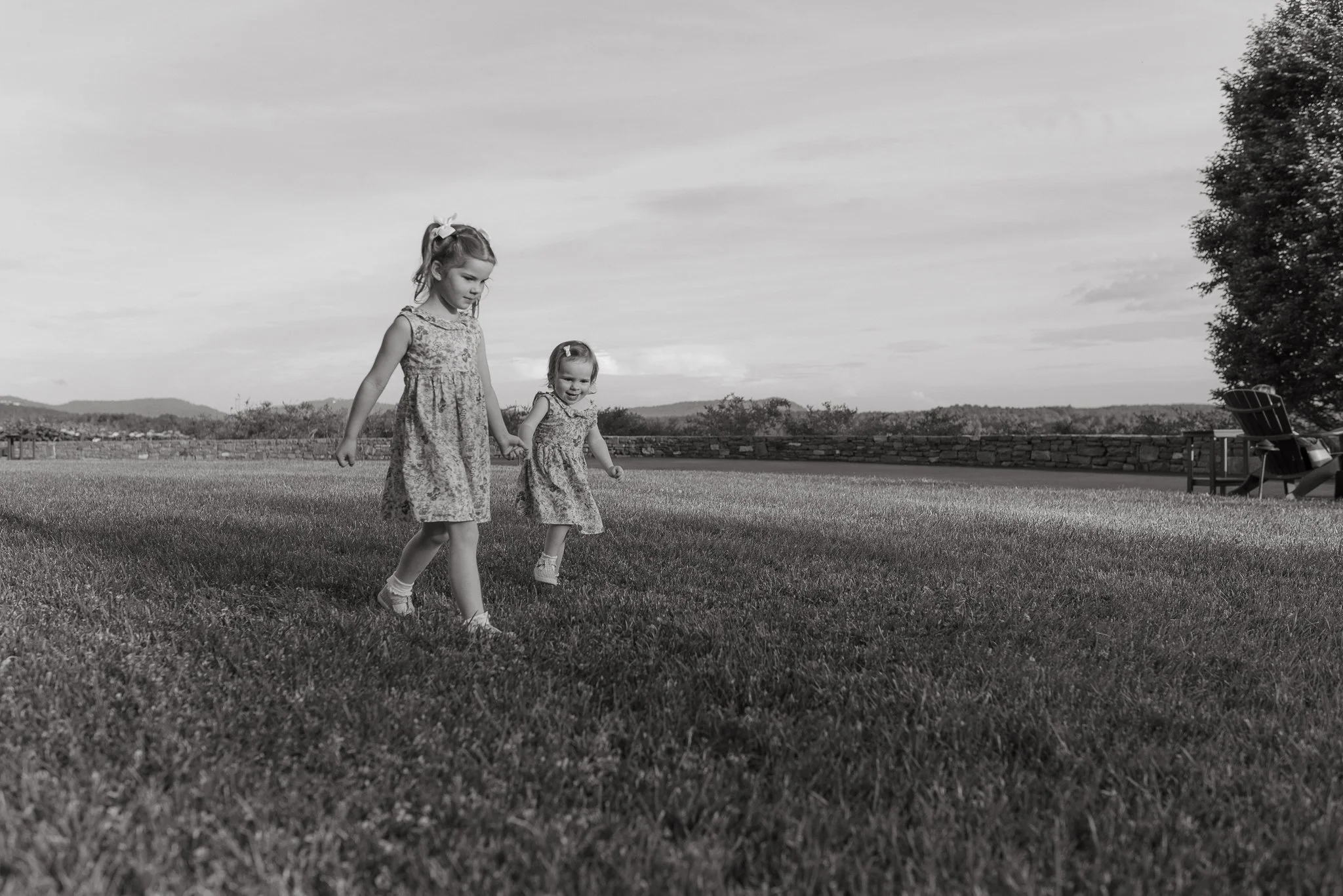 Two young girls wearing floral dresses walk hand in hand across a grassy field on a cloudy day, with trees, a stone wall, and outdoor chairs in the background.