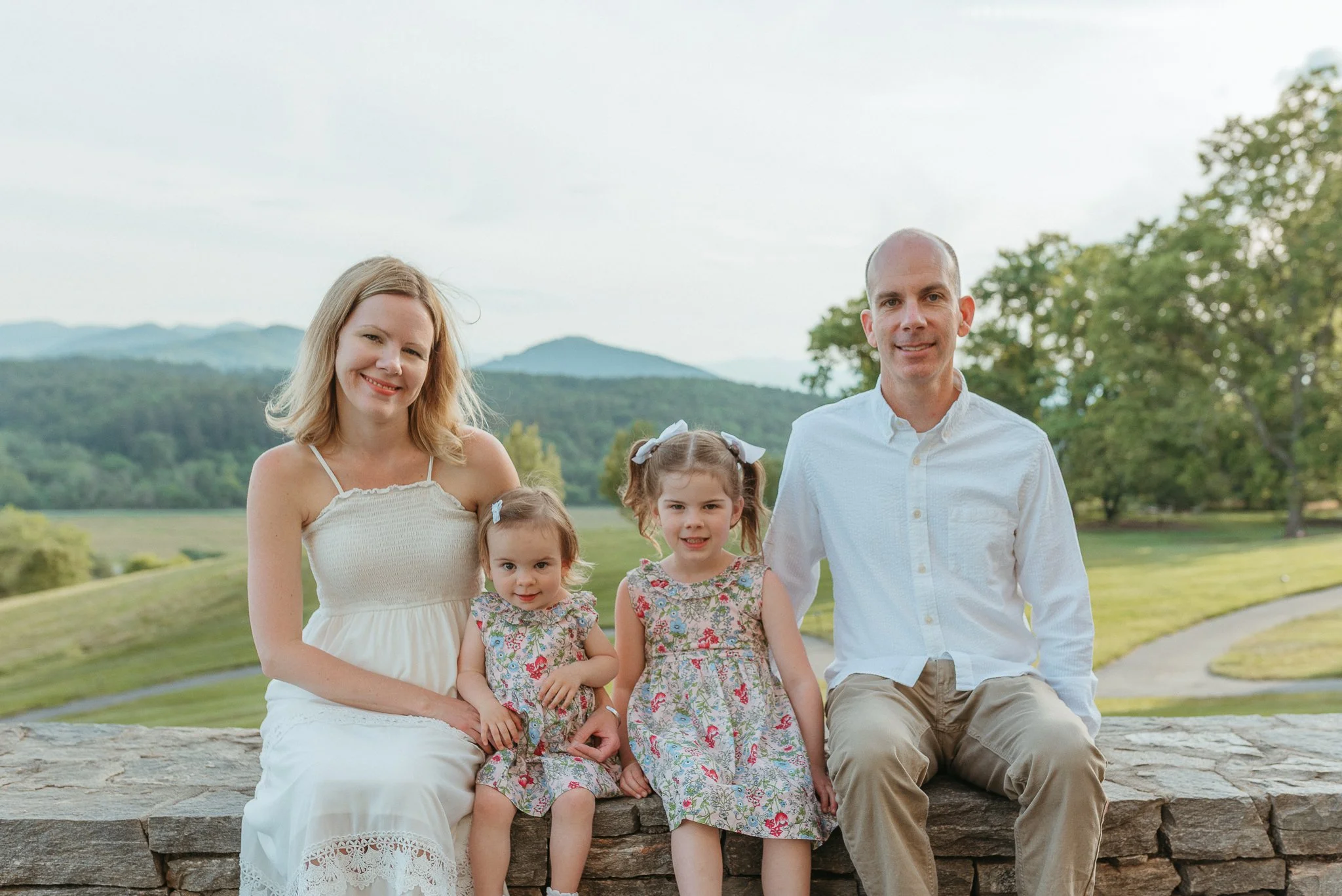 A family of five sitting on a stone wall outdoors, consisting of a mother, father, and three young girls, with trees and mountains in the background on a clear day.
