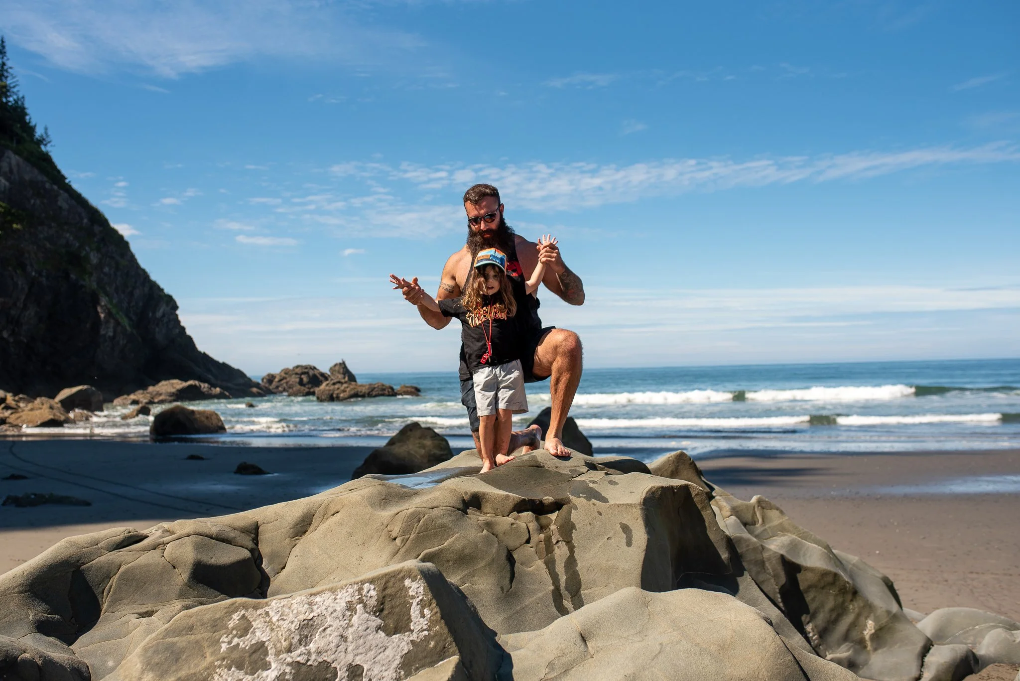 A man with a beard and sunglasses helping a young girl balance on a large rock on the beach. The sky is clear with some clouds, and the ocean waves are visible in the background.
