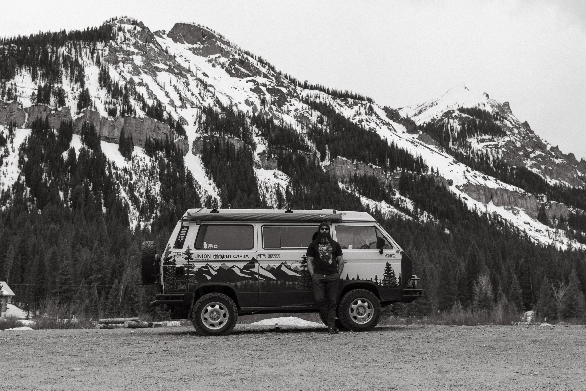 Black and white photo of a man standing next to a camper van in a mountainous, snowy landscape with pine trees and rugged peaks in the background.