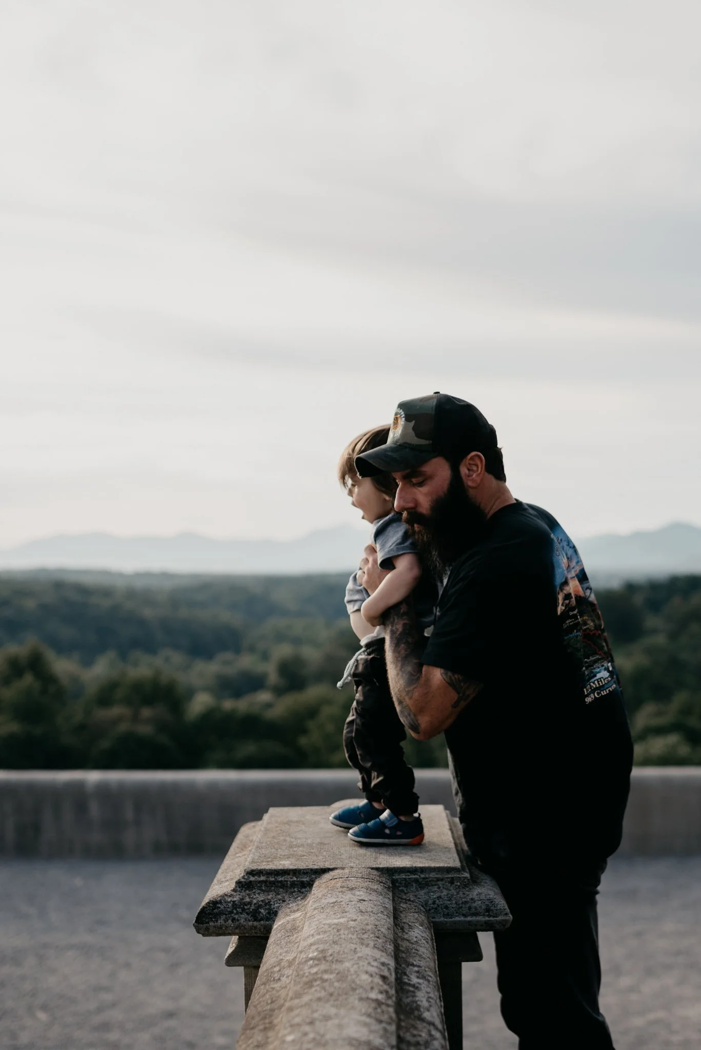 A bearded man wearing a cap and dark clothing is holding a child on a concrete railing, with the child standing on the railing. The man appears to be looking down while the child is looking forward. The background features a landscape with trees and distant mountains under a cloudy sky.
