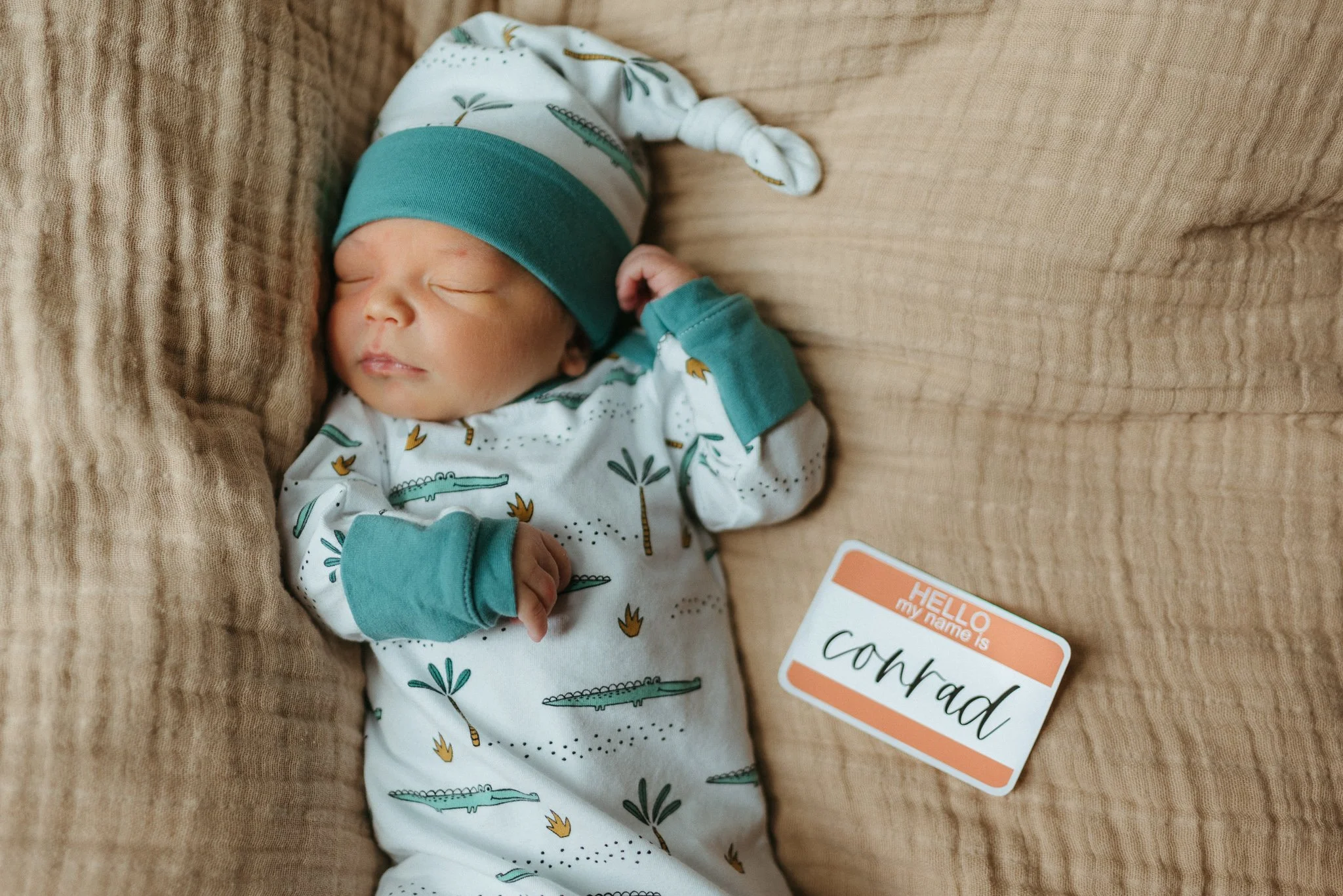 A sleeping baby dressed in a dinosaur patterned onesie and a teal and white hat, lying on a beige textured blanket with a "Congratulations" note next to them.