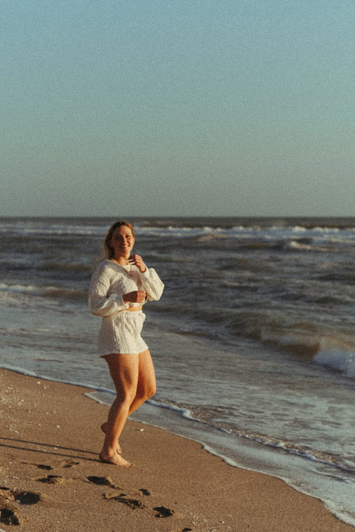 A woman in a white sweater and shorts walking along the beach at sunset, with the ocean and waves in the background.
