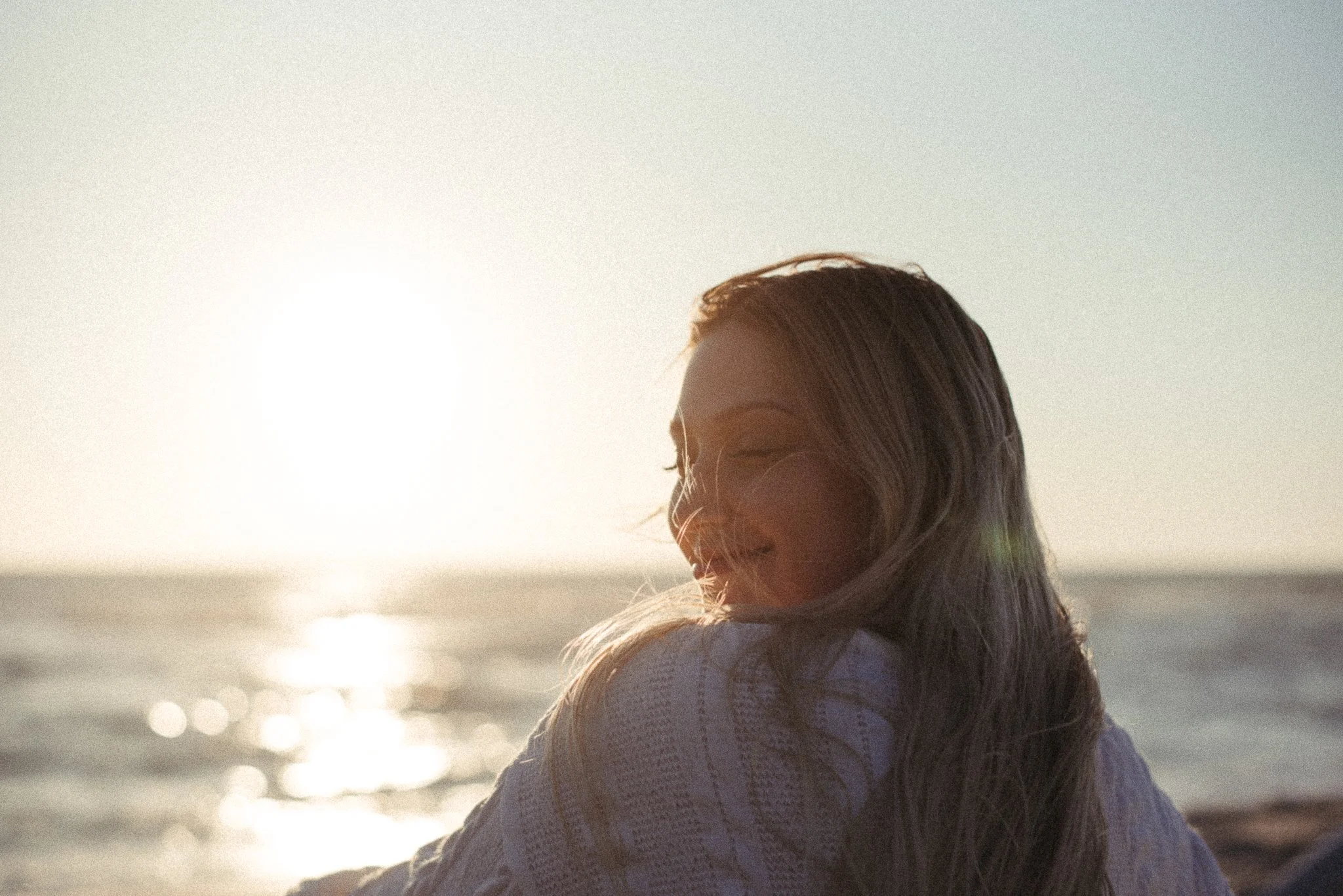 A woman with long hair smiling at the beach with sunlight reflecting on the water.