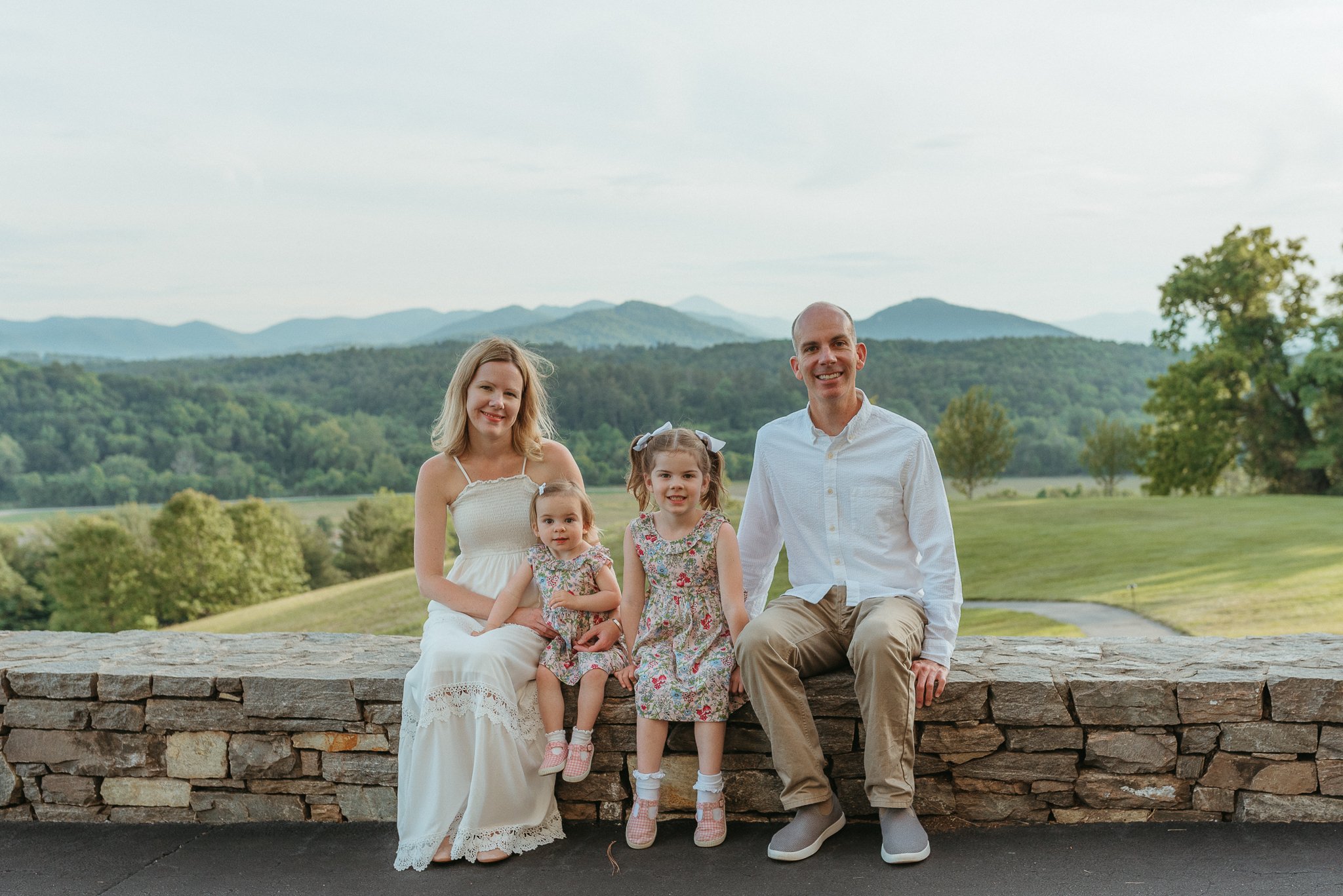 A family of five posing outdoors on a stone wall with lush green trees and mountains in the background, all smiling at the camera.