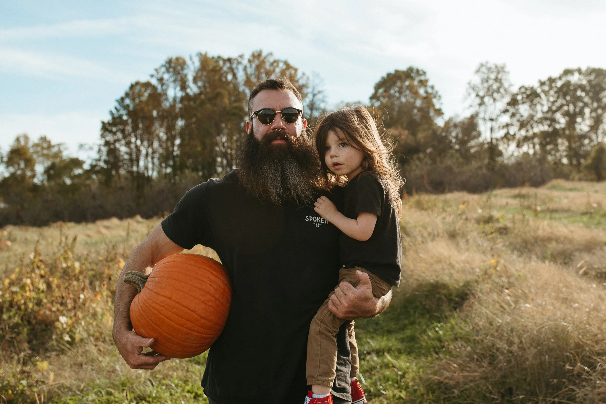 A bearded man with sunglasses holding a young girl while standing outdoors in a field with trees in the background. The man is holding a pumpkin.