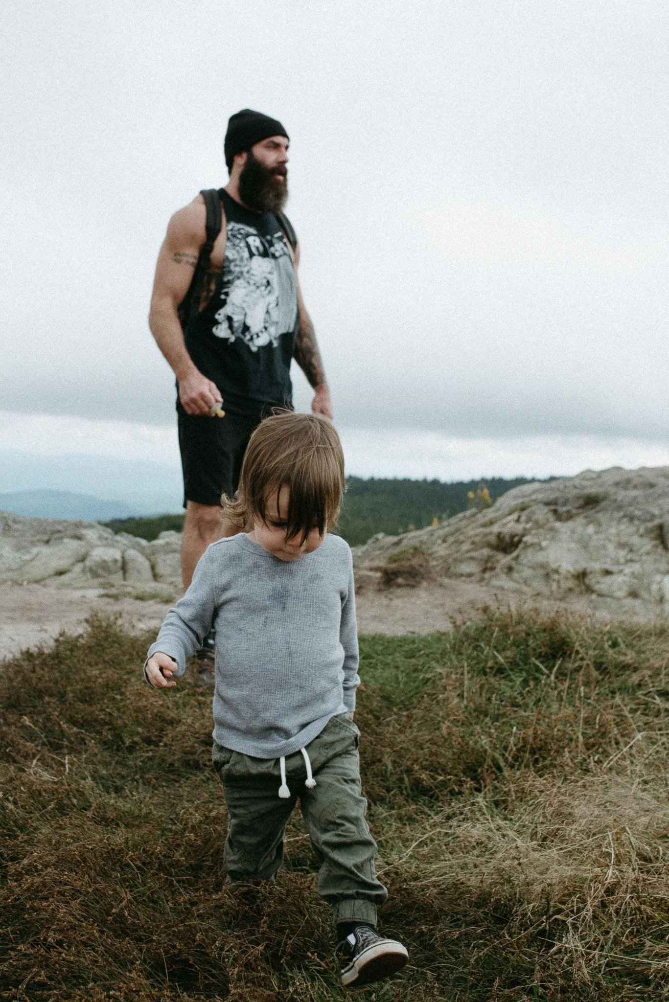 A man with a beard, wearing a black beanie, sleeveless shirt, and backpack, walking outdoors on a rocky terrain with a young boy in front of him, in an open natural landscape under a cloudy sky.
