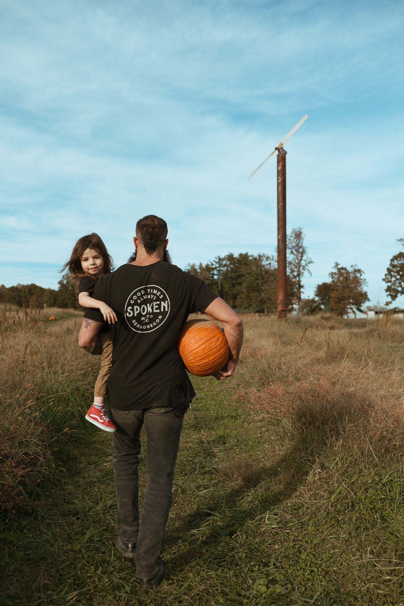 A man carrying a young girl on his arm as they walk on a grassy path in a field. The man holds a pumpkin under his arm. The girl is wearing red shoes. There are trees and a tall pole with a windmill blade attached in the background, under a partly cl