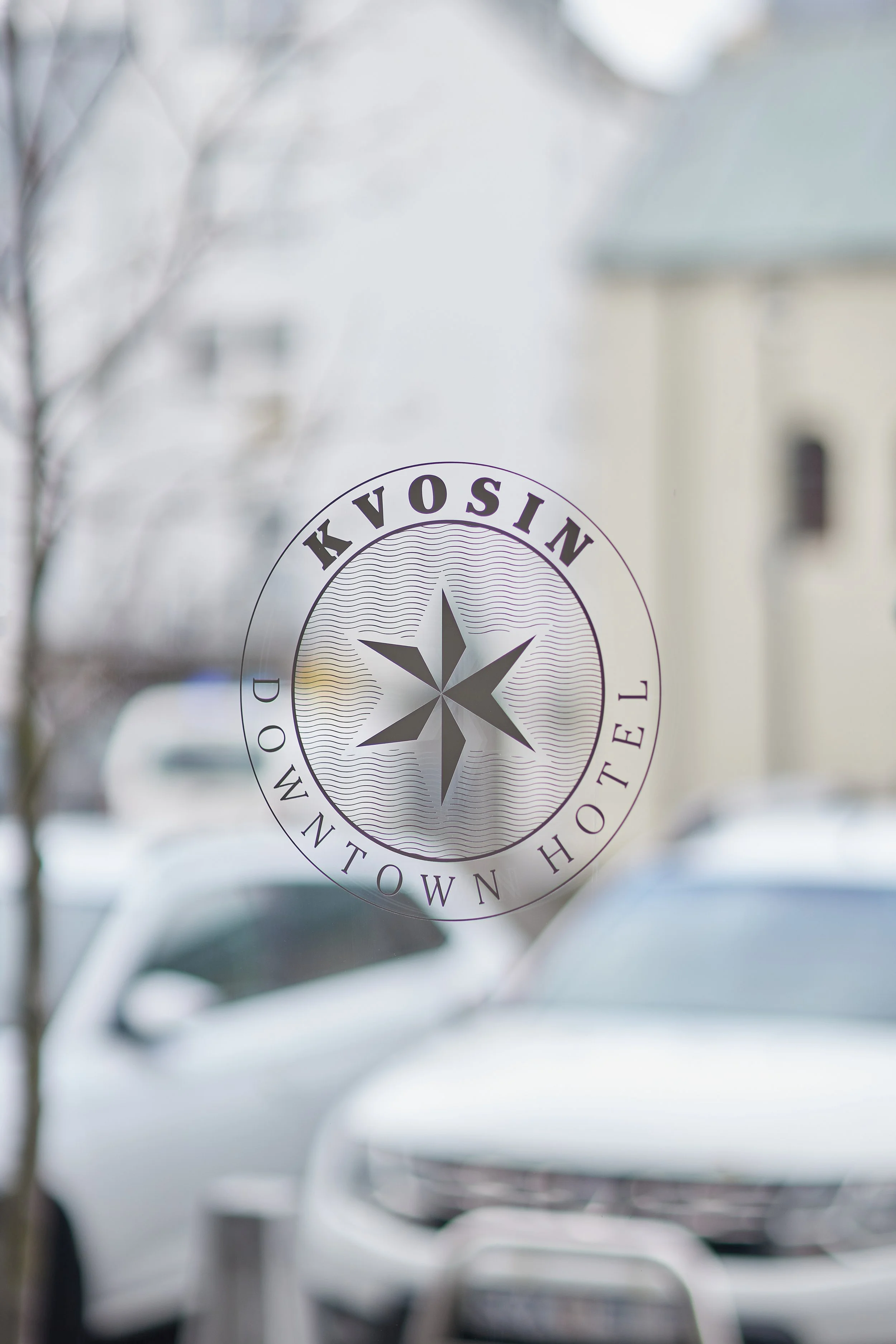 Close-up of a glass window or door with a circular logo for a company named 'Kvosin Hostel' and a compass symbol, with a reflection of white cars parked outside and a blurred tree branch.