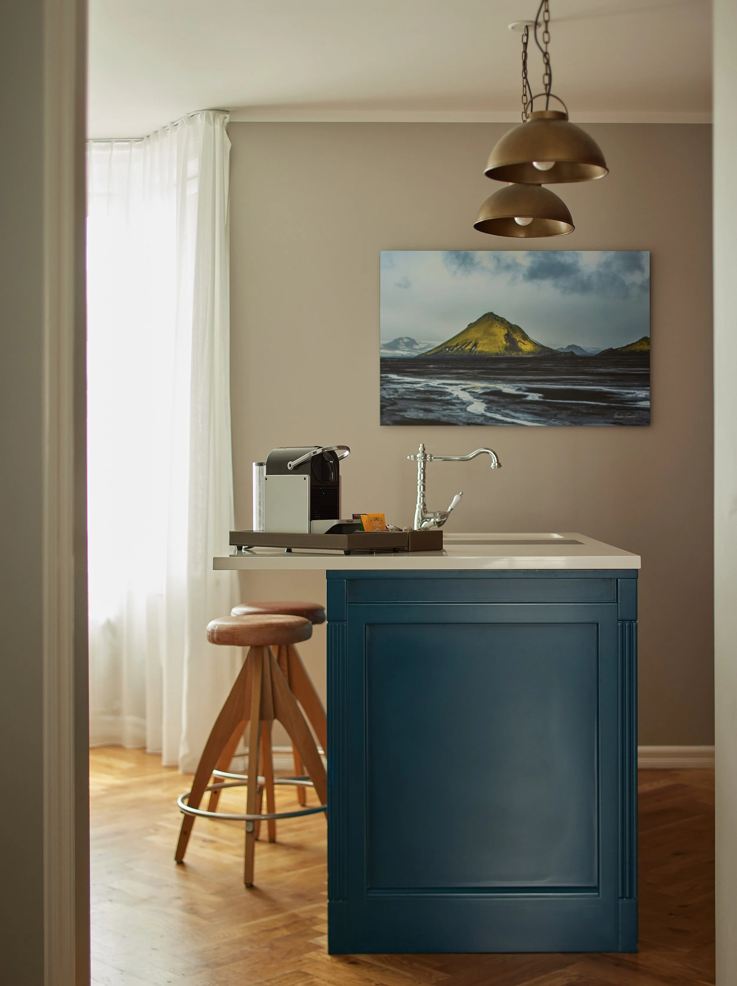 A kitchen island painted in dark teal with a white countertop, a small coffee machine, and a small yellow box. Two wooden bar stools with circular seats are beside it. In the background, there's a wall with a landscape painting of a volcano and a win