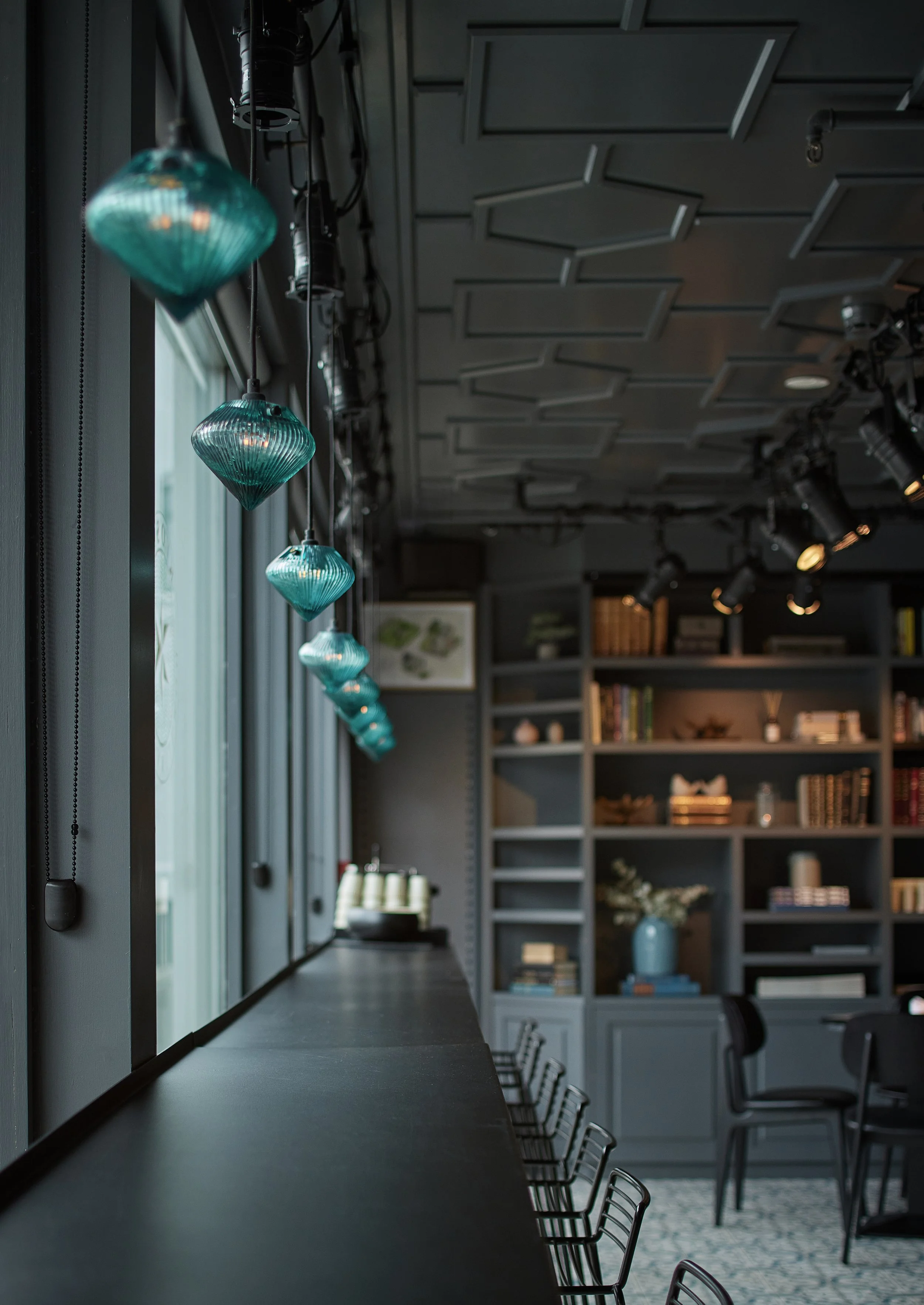 A row of turquoise pendant lights hanging beside a large window in a modern interior dining or lounge area, with shelves full of books and decorative items in the background.