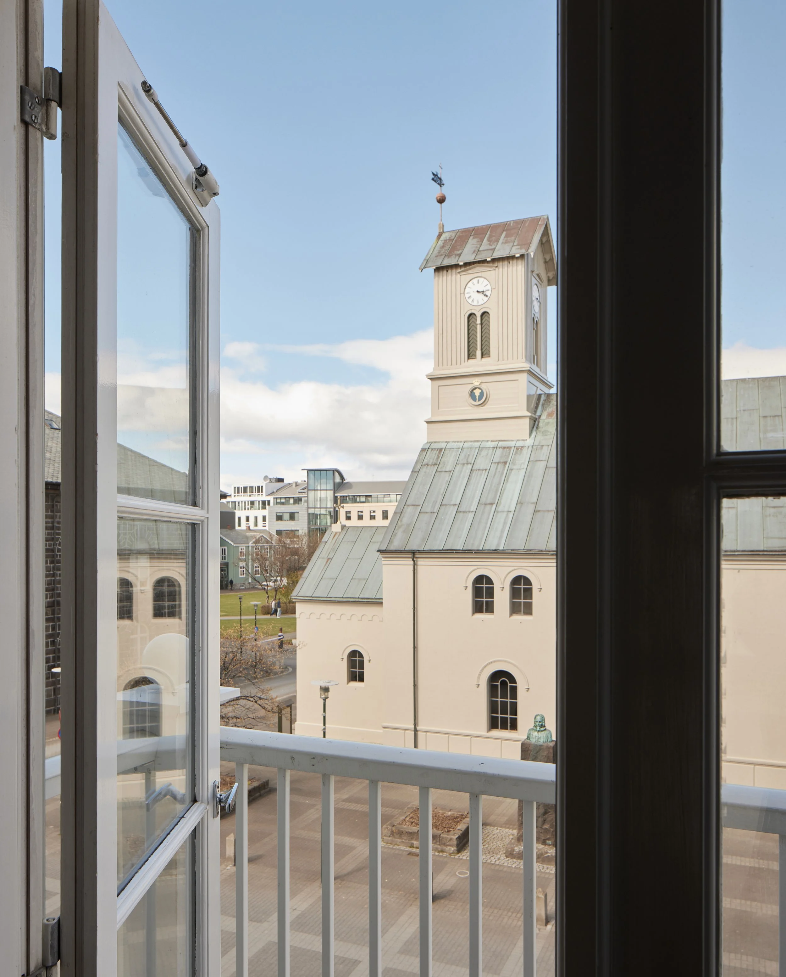 View from a balcony showing an old building with a clock tower and a weather vane on top, with modern buildings in the background under a partly cloudy sky.