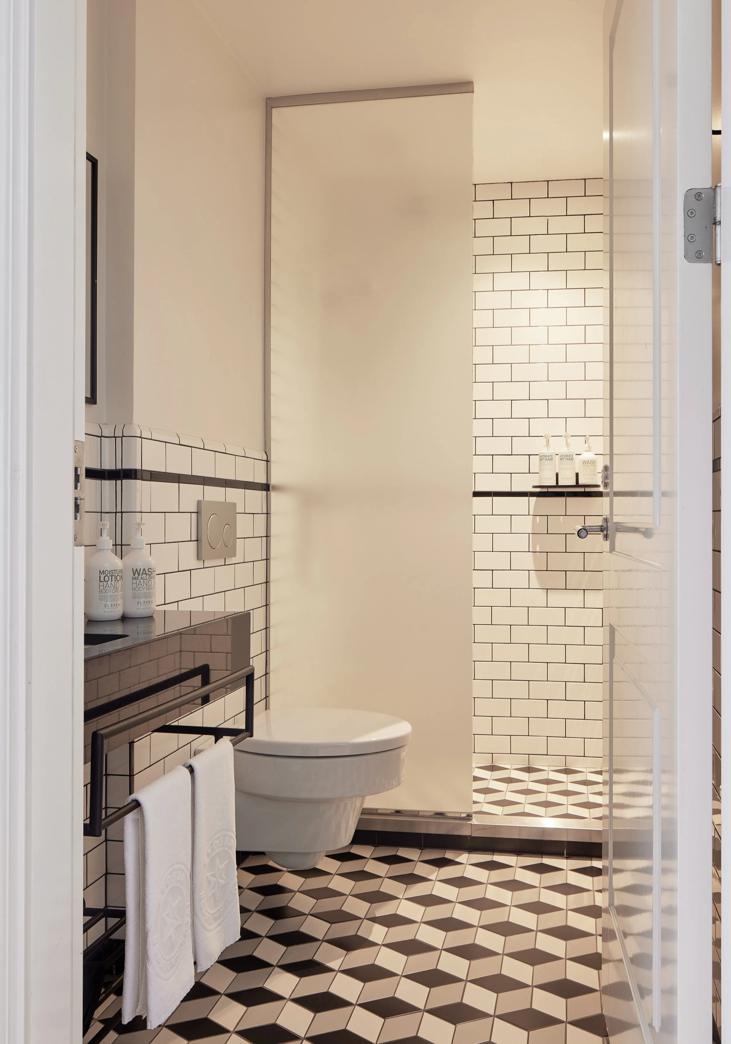 Modern bathroom with black and white geometric patterned floor tiles, white subway wall tiles, a floating toilet, a towel rack with white towels, and shelves with toiletries.