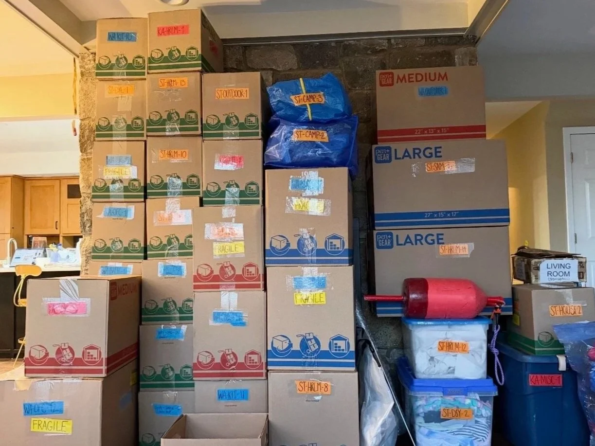 Stacked moving boxes labeled with various room names and contents, some sealed and others open, with a blue bag on top and a red propane tank nearby, indicating storage or moving in process.
