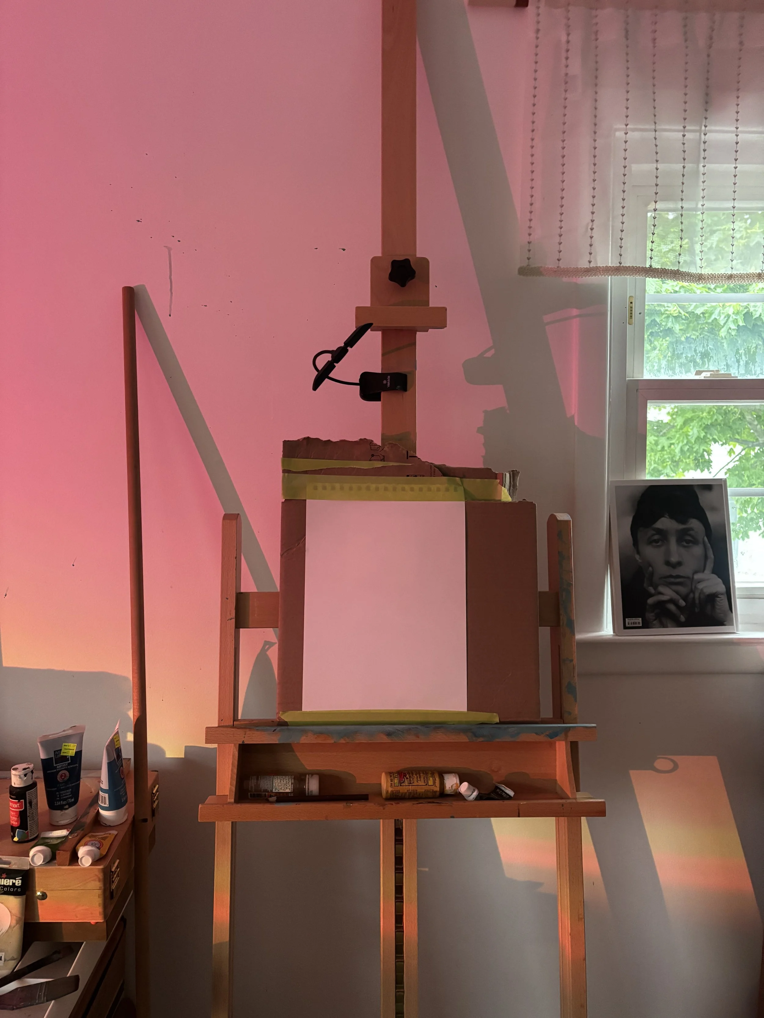 A wooden art easel holding a blank canvas in a room with a window. The window has sheer curtains, and a person's face is visible in a black-and-white photograph near the window. Various art supplies are on shelves below the easel and on the left side of the image.