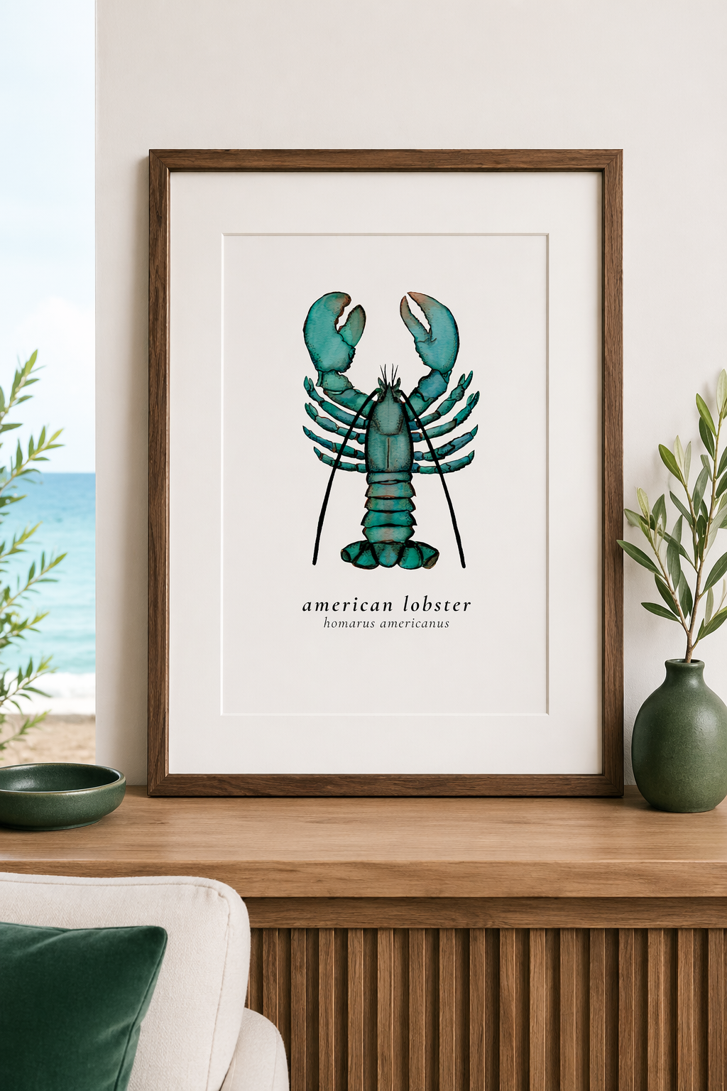 A framed botanical print of an American lobster (Homarus americanus) on a wall, with green plants and decor on a wooden sideboard, beach visible through a window on the left.