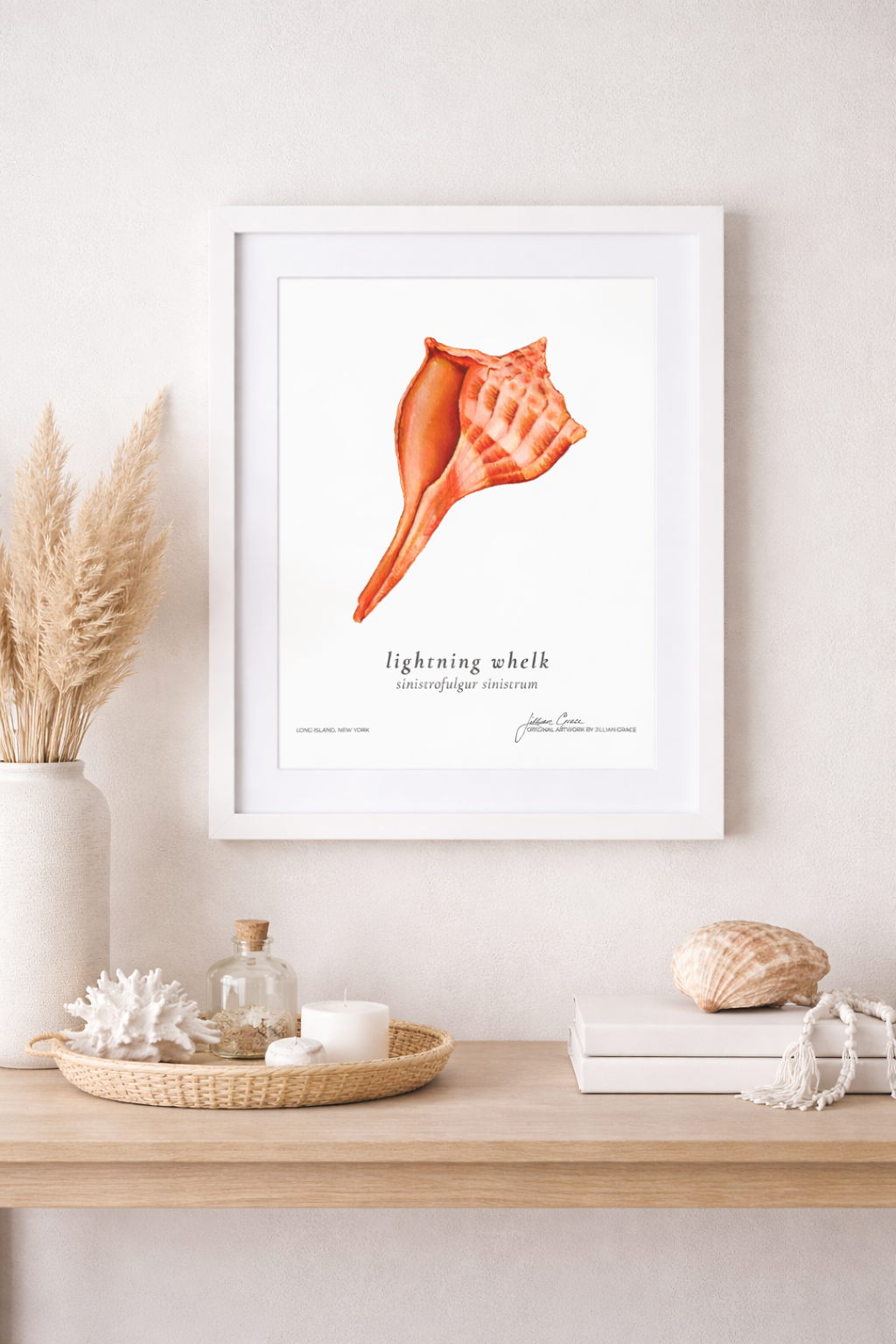A white framed art print of a pink conch shell on a white wall, with dried pampas grass in a white vase, a wicker tray with coral, a small glass bottle, and candles, and a shell and books on a light wooden shelf.