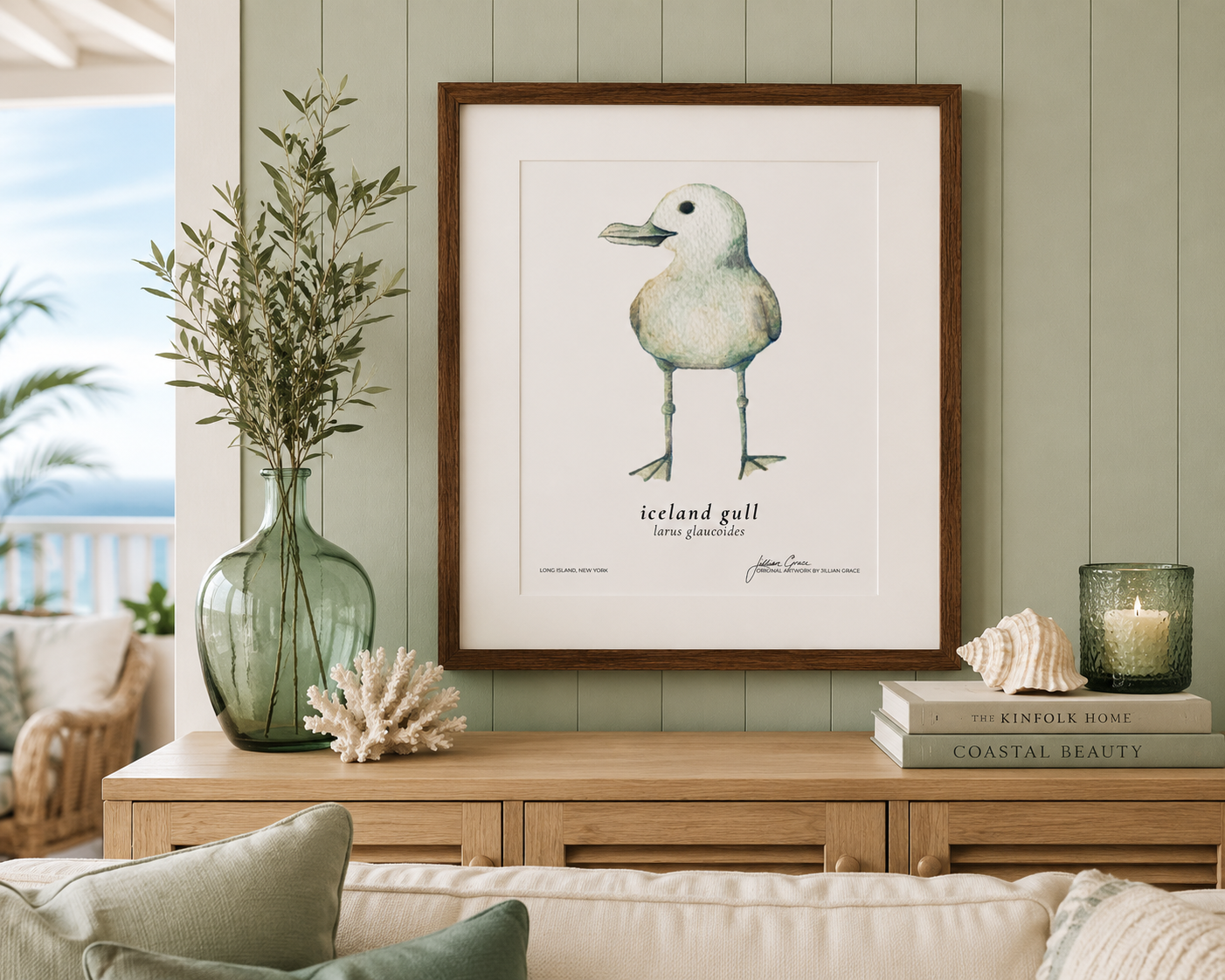 Decorative coastal-themed room with a framed illustration of an Iceland gull on a green wall, a glass vase with greenery, a coral ornament, books, and a lit candle in a glass holder on a wooden sideboard.