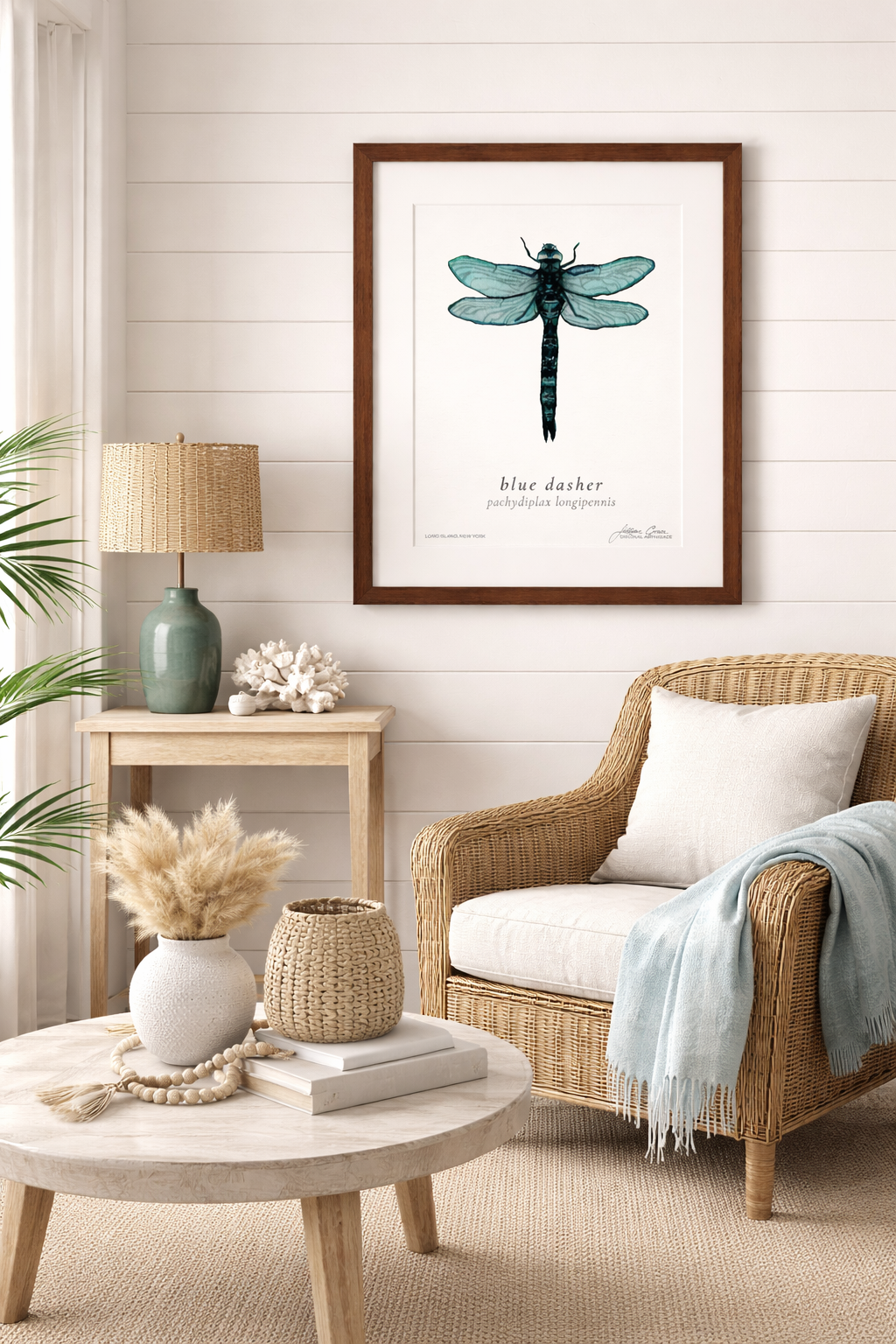 A living room with a framed illustration of a blue dragonfly on a white paneled wall, a wicker armchair with a white cushion, a light blue throw blanket, a small wooden side table with a green ceramic lamp, coral, and white decorative items, a round wooden coffee table with a white textured vase with dried grasses, a beige woven basket, white books, and a beaded necklace.