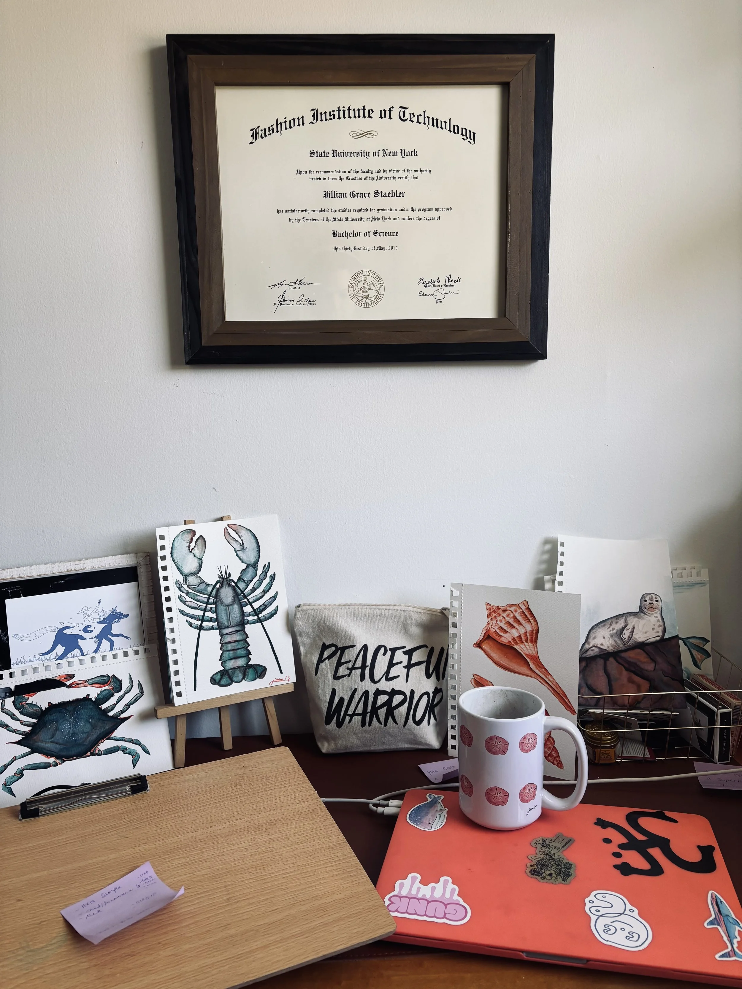 A framed certificate from the Fashion Institute of Technology hangs on a white wall. On the table below, there are various art prints, a white mug with pink flower patterns, and a red laptop covered with stickers, including a pink sticker with text, a skull, and a rainbow-colored brain.