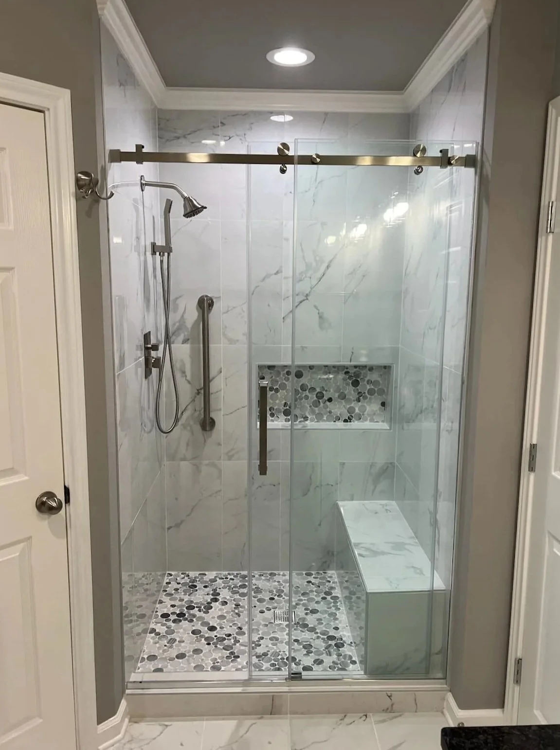 A modern walk-in shower with glass doors, marble tile walls, a built-in niche with pebble tile, a shower bench, and two showerheads in a residential bathroom.