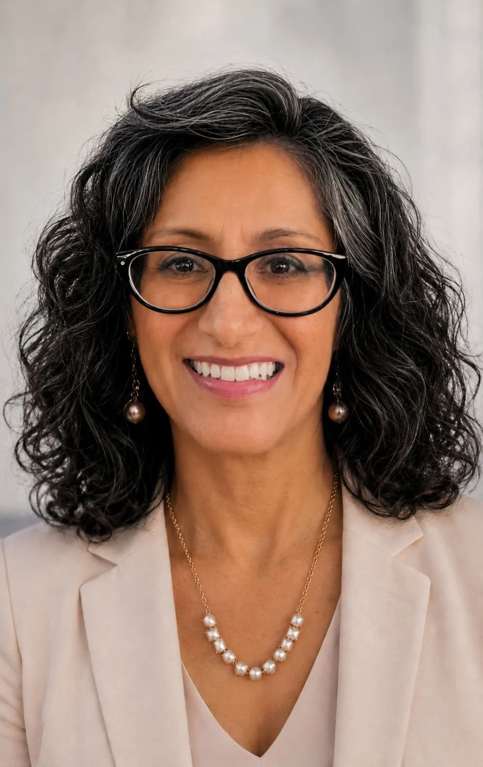 Close-up of a smiling woman with curly dark hair, wearing glasses, pearl earrings, a pearl necklace, and a beige blazer.