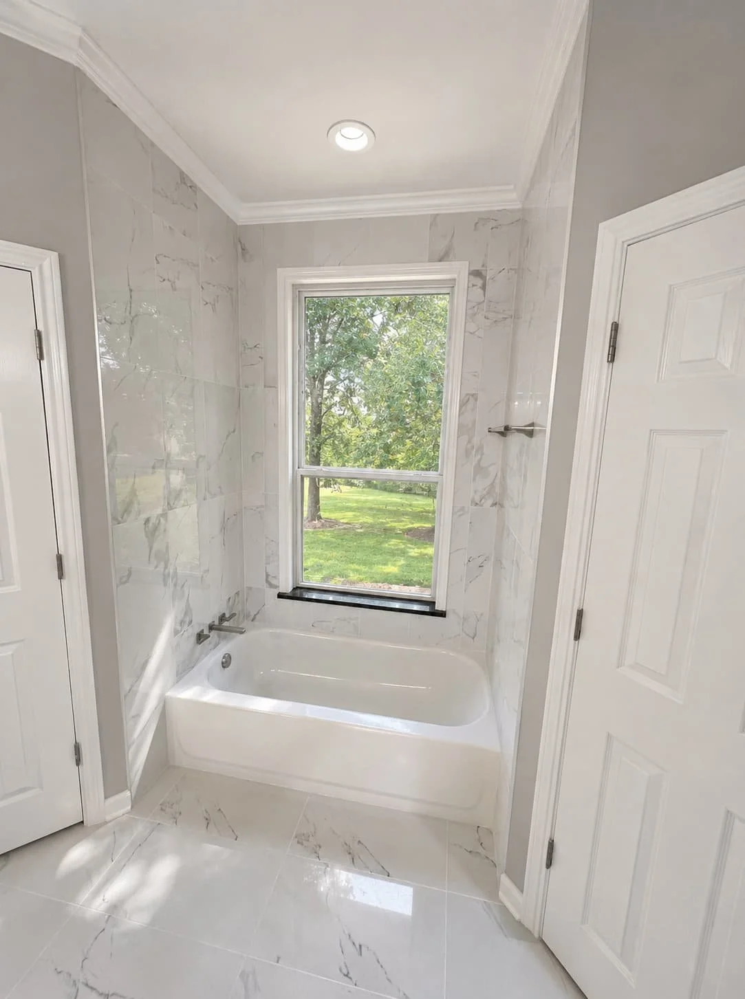 Bathroom with a large window, bathtub, marble wall tiles, and white doors.