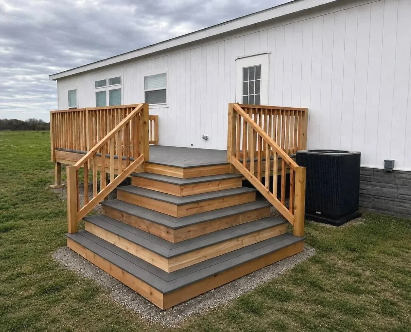 A wooden deck with stairs leading up to it, attached to a white building. The deck has wooden railings and a gray surface, with an air conditioning unit nearby. The background shows a grassy field and an overcast sky.